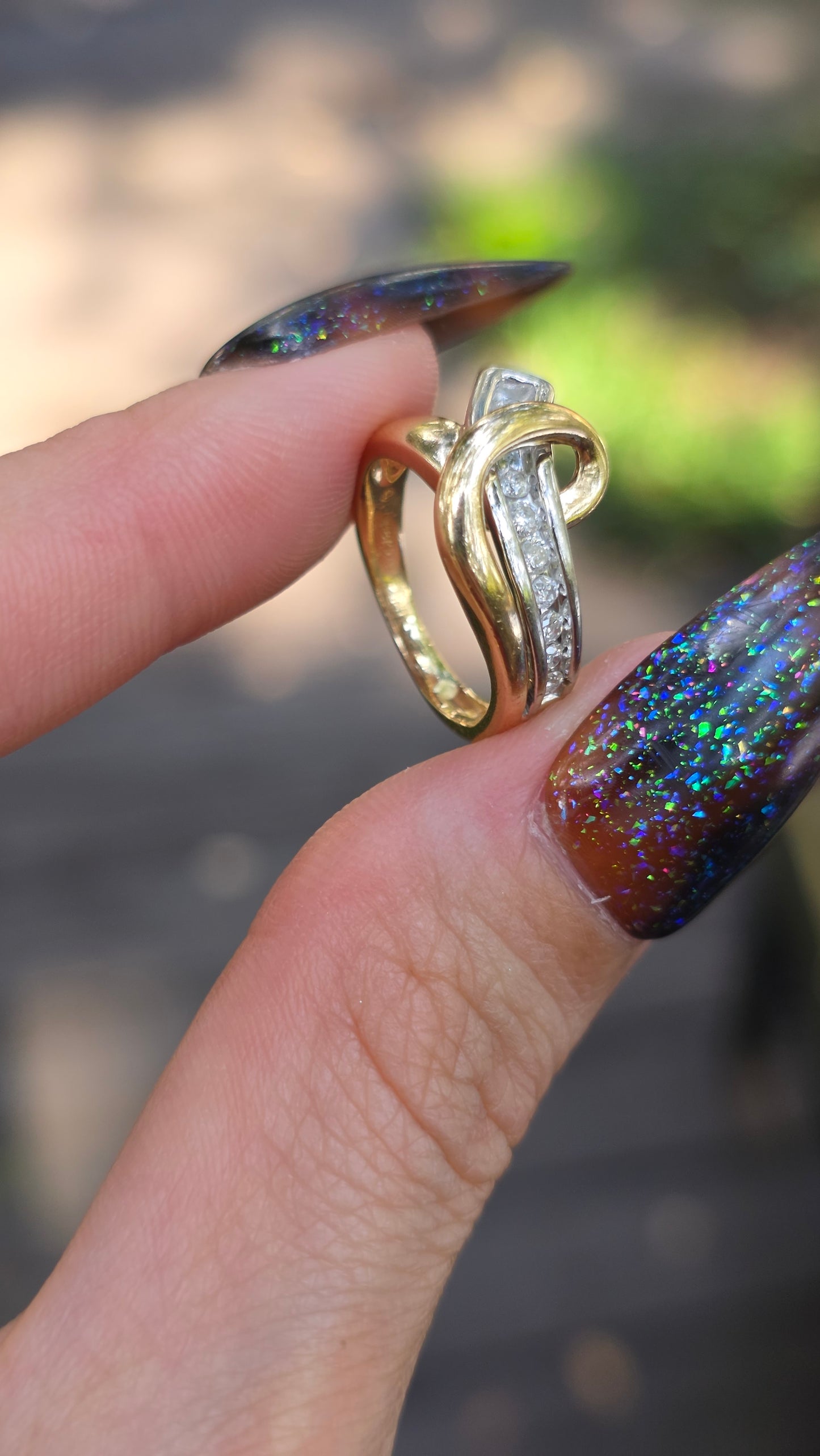 A close-up of a hand with glittery dark nail polish holding a gold ring with a curved band and a row of small clear stones.