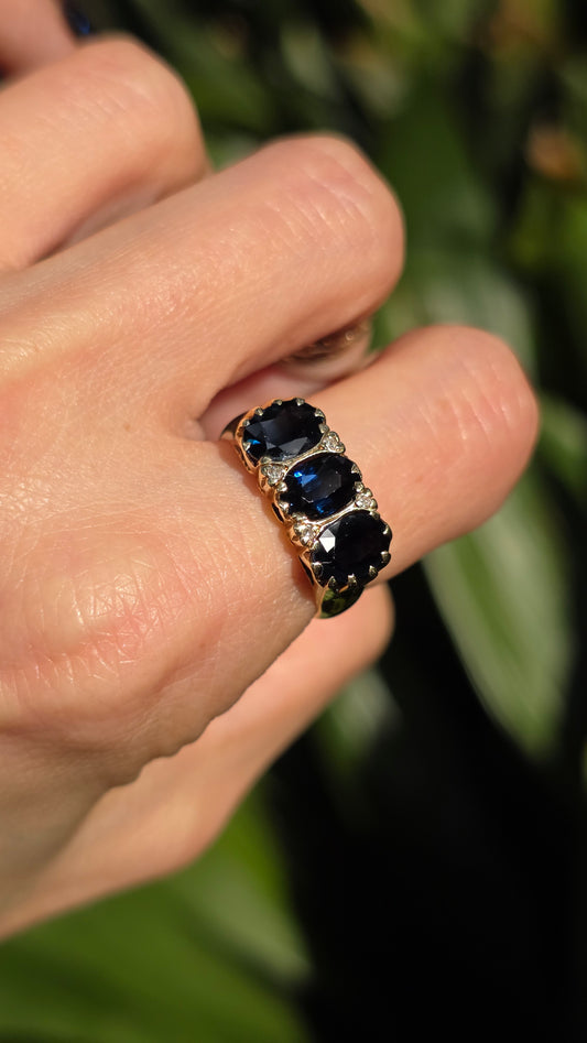 A close-up of a hand wearing a vintage-style gold ring with three oval-cut dark blue sapphires and small clear accent stones, set against a blurred green background.