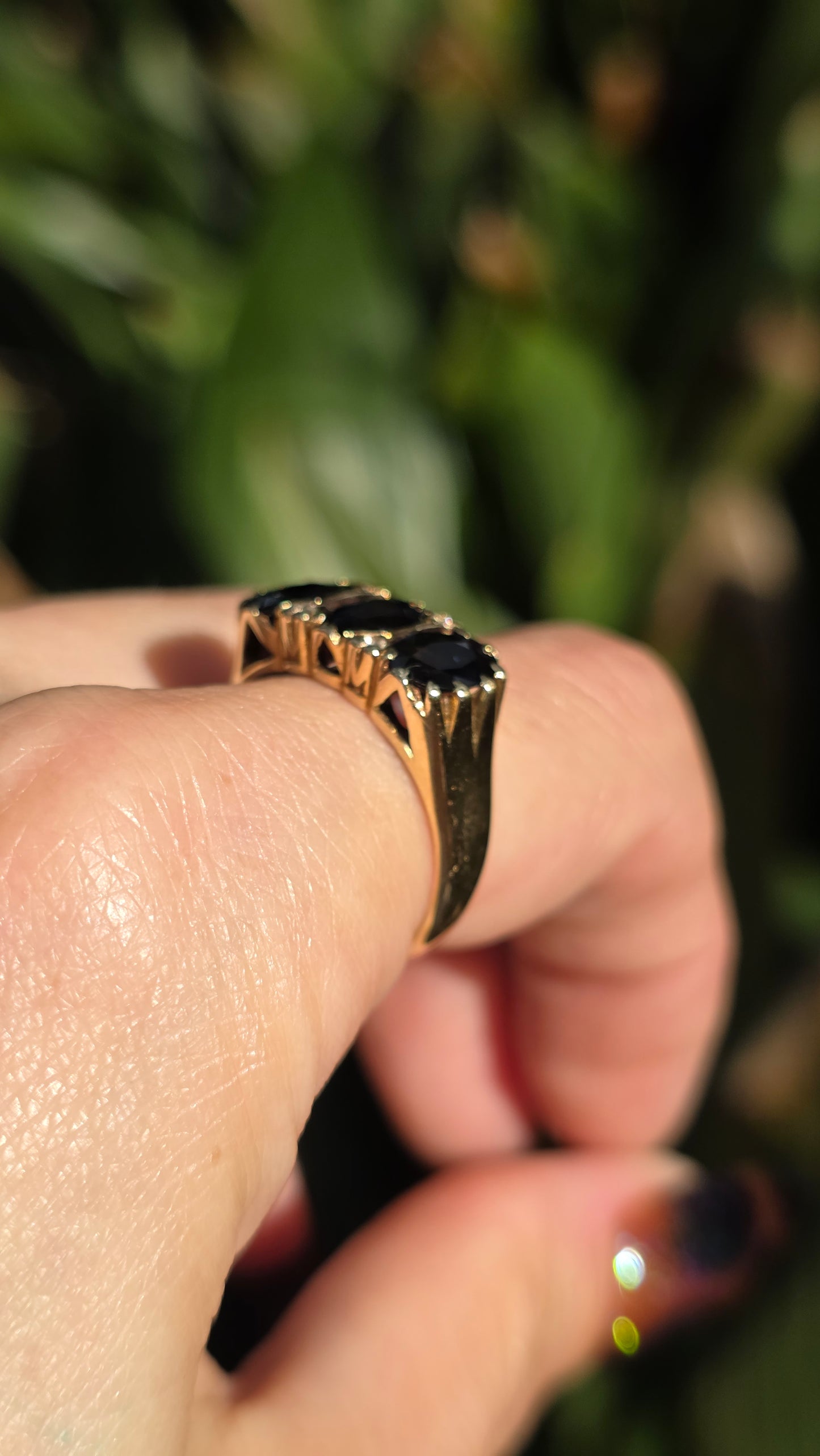 A gold ring with three black oval stones worn on a finger, shown in a close-up shot against a blurred green background.