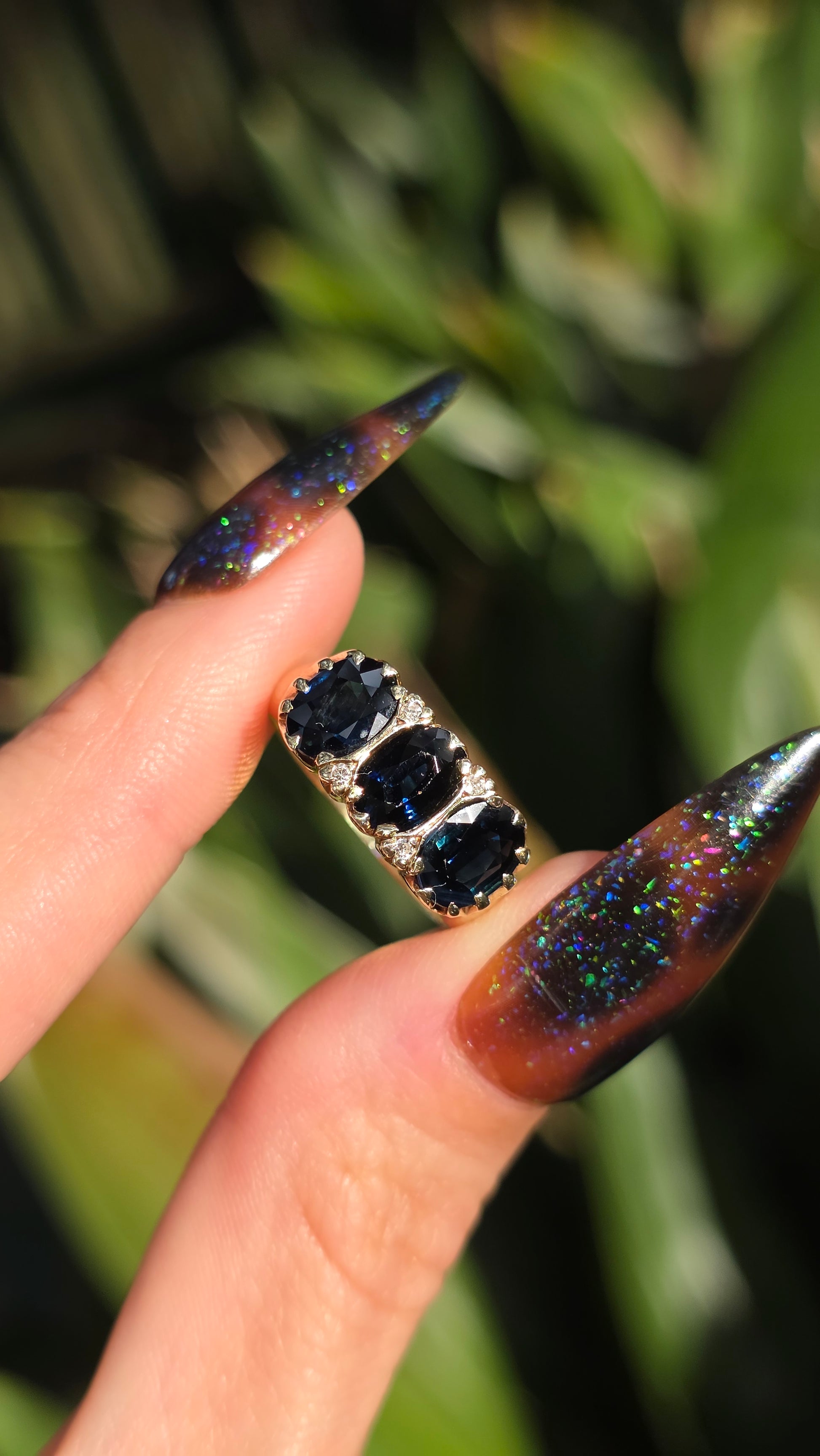 A close-up of a hand with long, sharp stiletto nails, showcasing a dark iridescent glitter nail design on one finger and a silver ring with three round black gemstones on the other, set against a blurred green background.