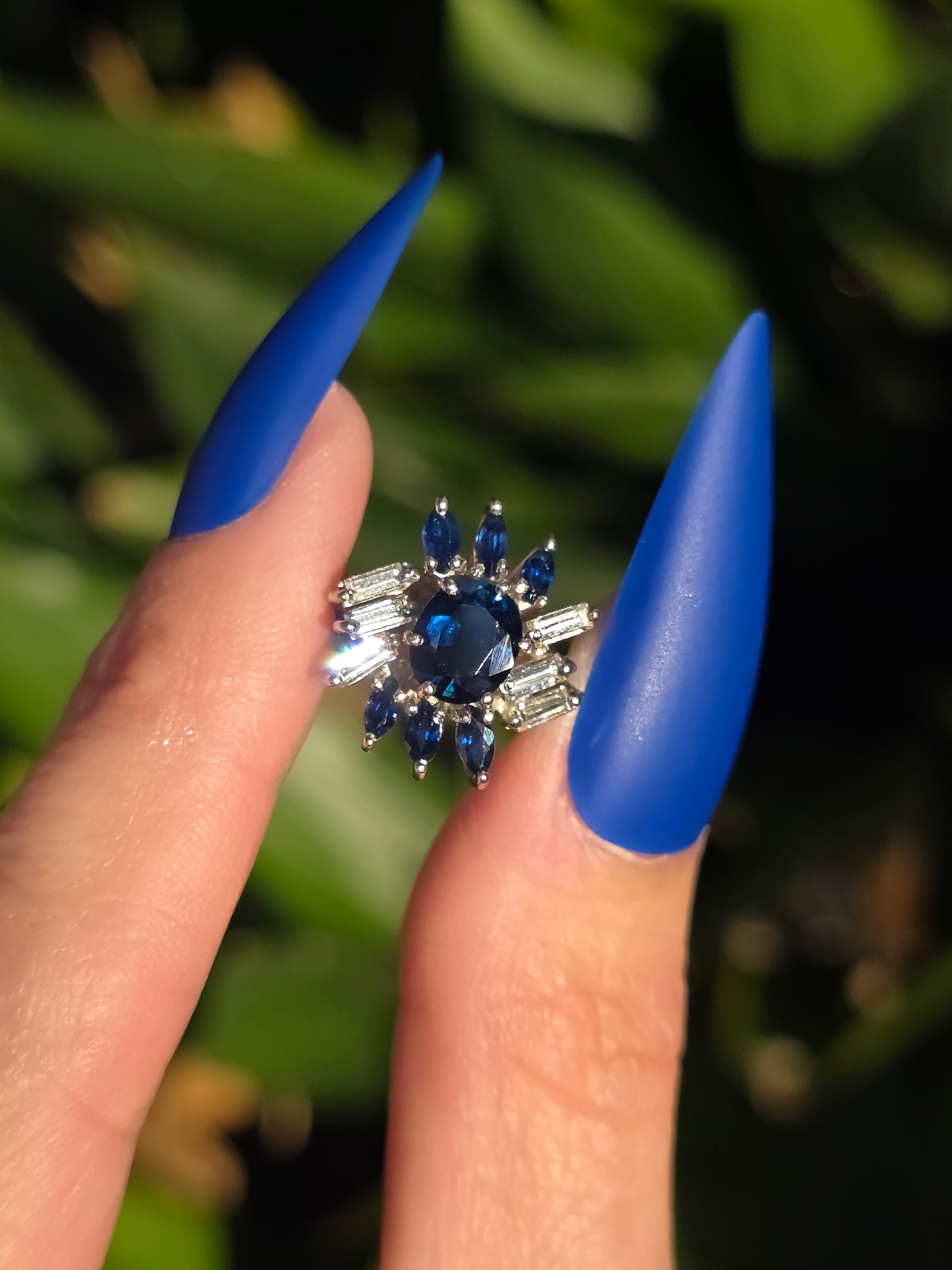 Close-up of a hand holding a ring with blue gemstones against a blurred green background