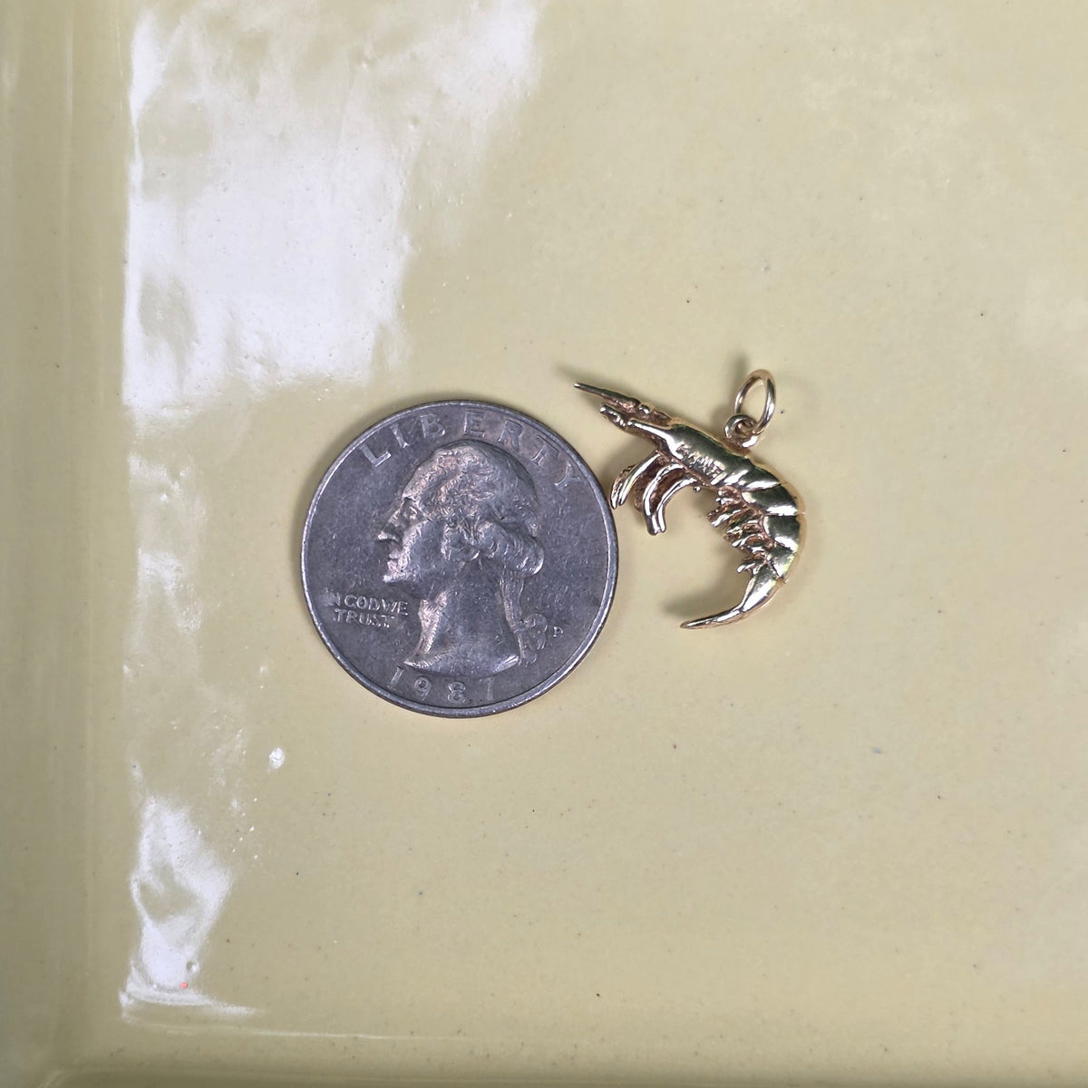 A small gold fish charm displayed next to a US quarter coin on a glossy pale yellow ceramic dish, with a dark wood surface visible at the bottom edge.