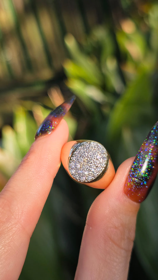 A close-up of a hand holding a gold ring with a round, heavily textured silver pavé face, set against a blurred green foliage background.