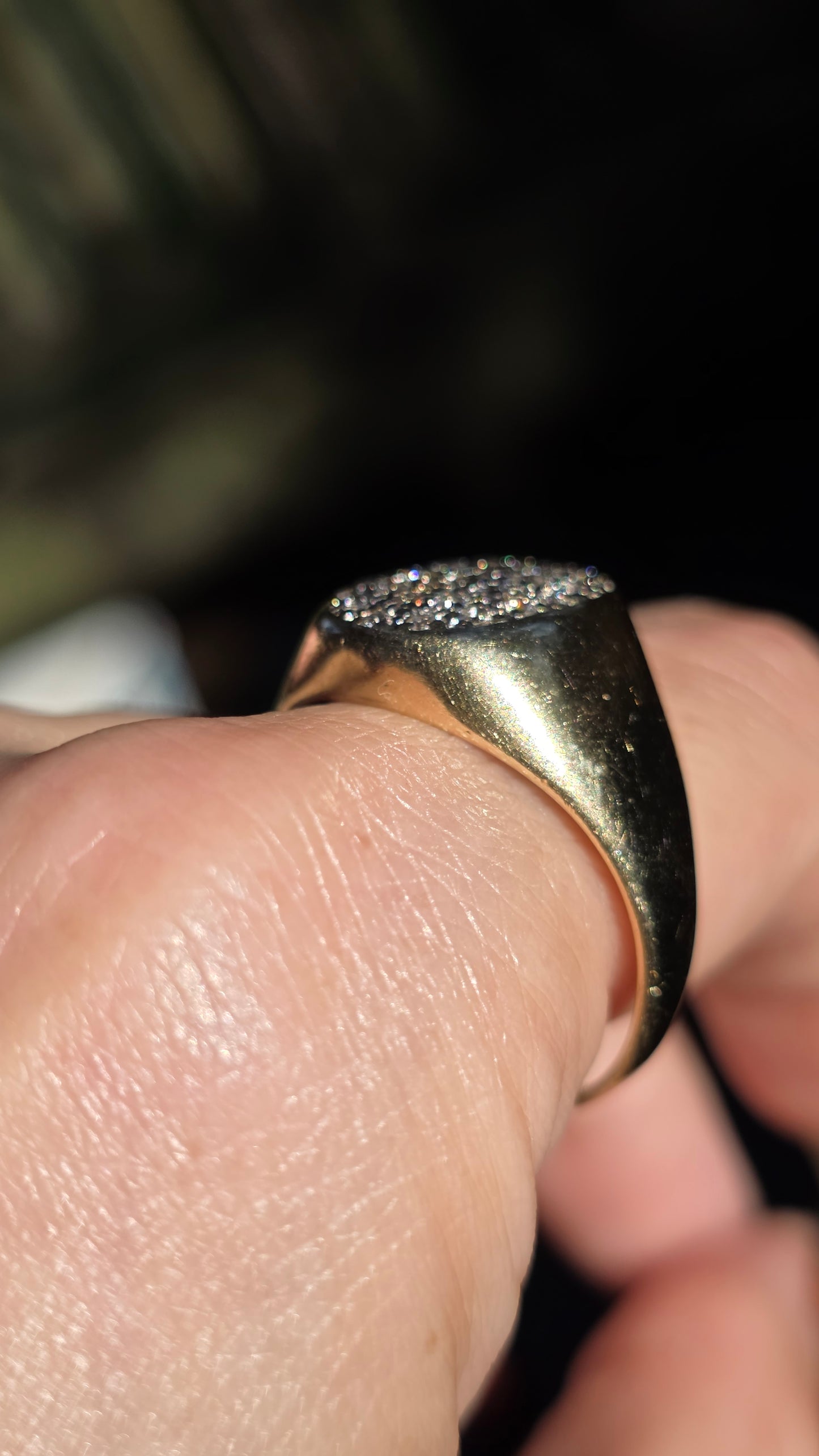 A close-up of a gold signet ring with a textured, diamond-like paved setting, worn on a finger against a dark, out-of-focus background.