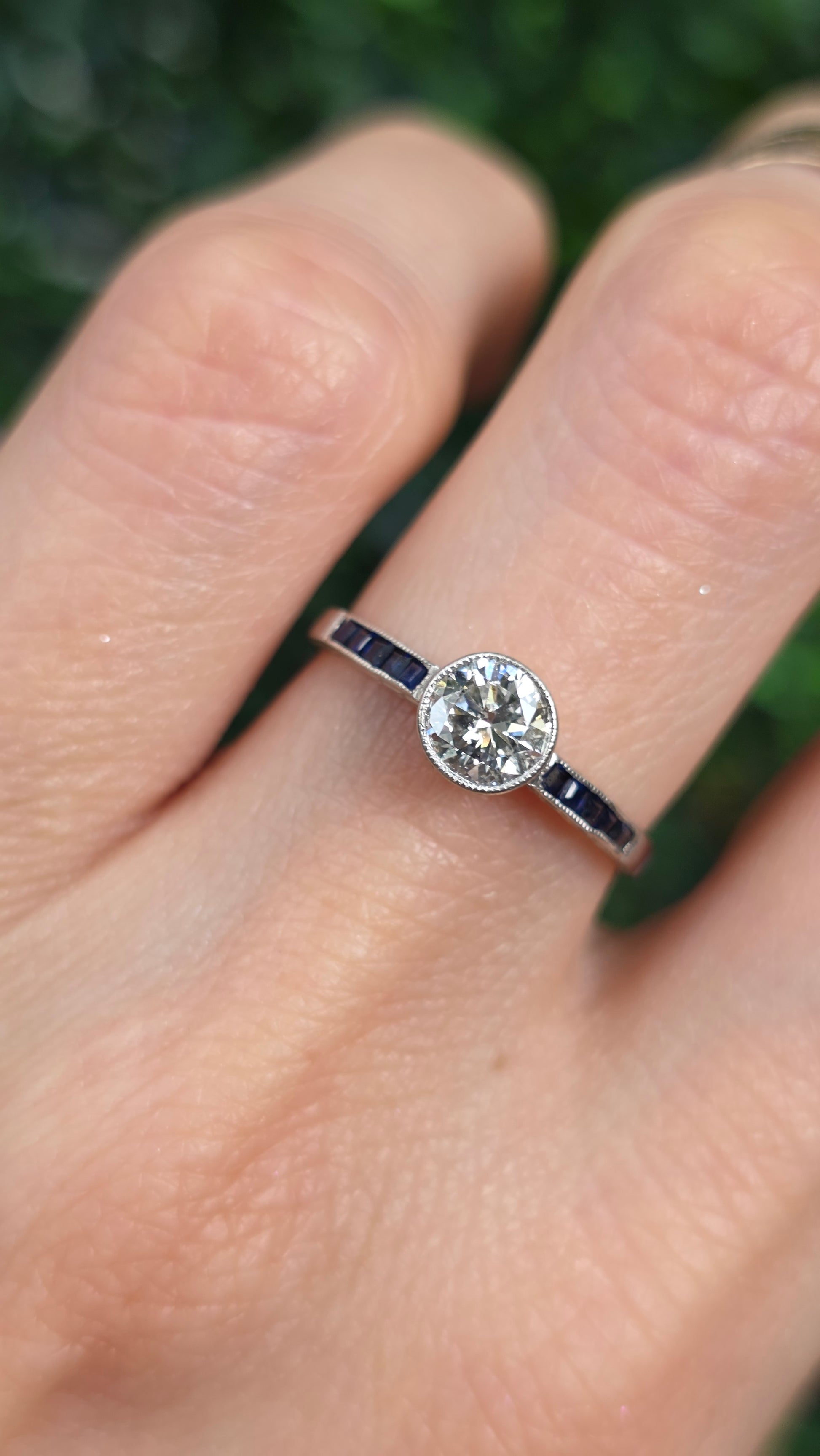 A close-up of a hand wearing a silver-toned ring with a round clear gemstone and small dark accent stones on the band, set against a blurred green outdoor background.