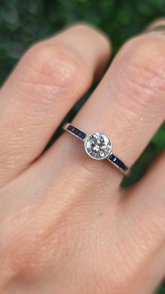 A close-up of a hand wearing a silver-toned ring with a round clear gemstone and small dark accent stones on the band, set against a blurred green outdoor background.
