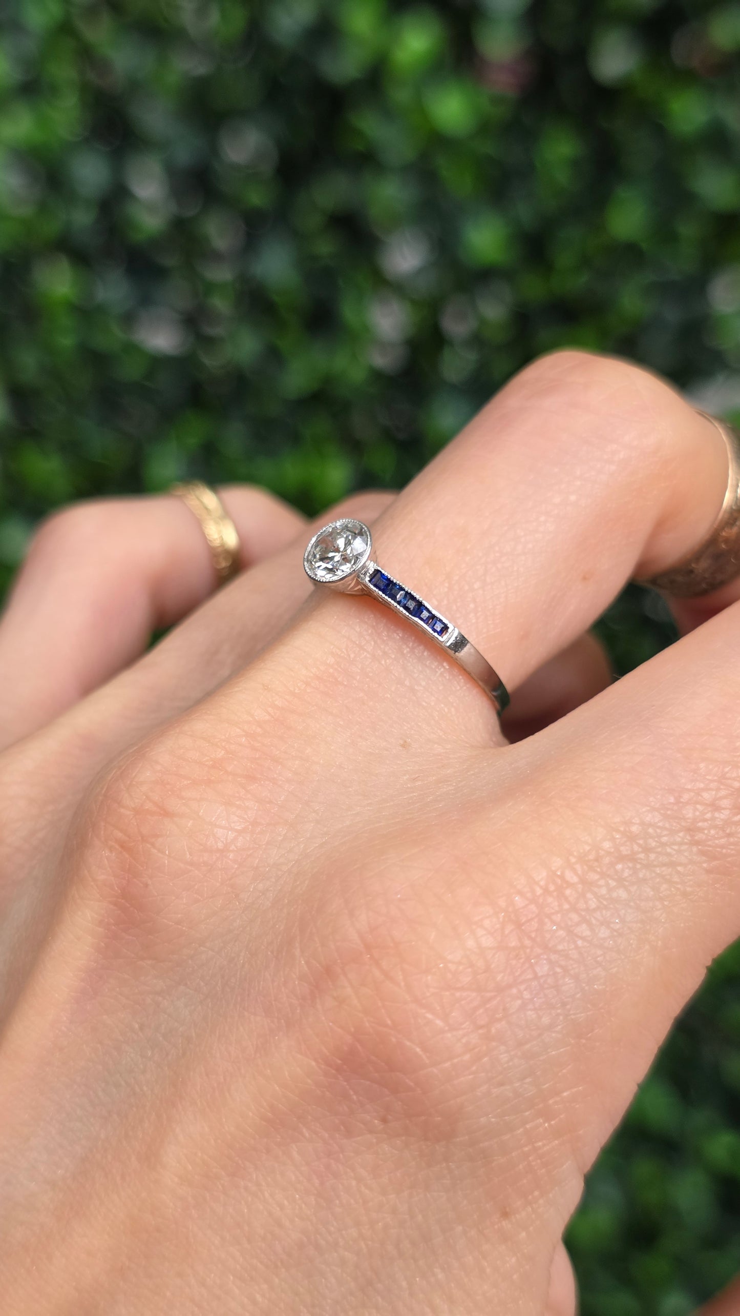 A close-up of a hand wearing a silver ring with a round clear gemstone and a small inlaid blue accent on the band, set against a blurred green hedge background.