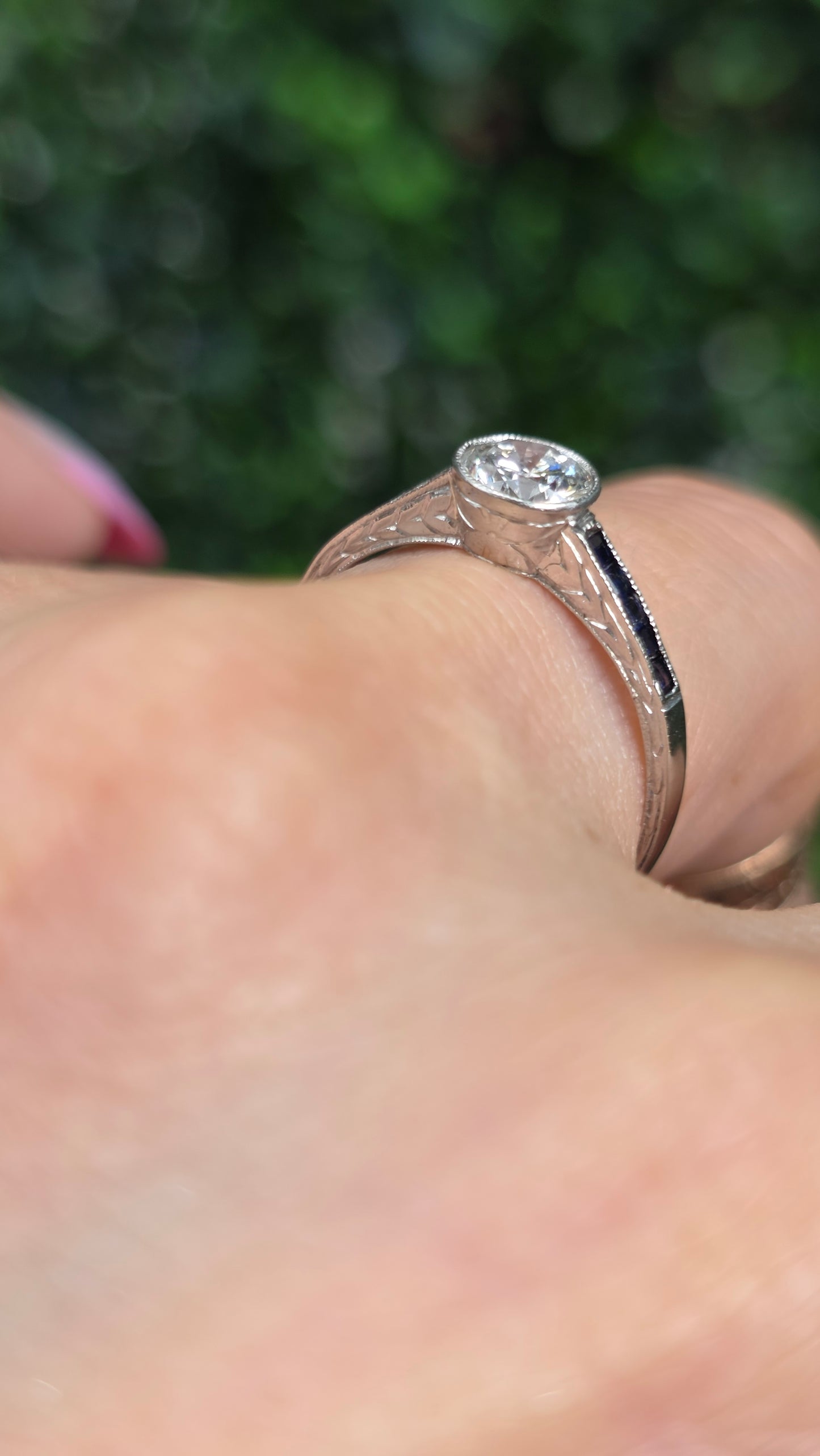 A close-up shot of a hand wearing a vintage-style rose gold ring with an oval clear gemstone and a black inlay on the band, set against a blurred green background.
