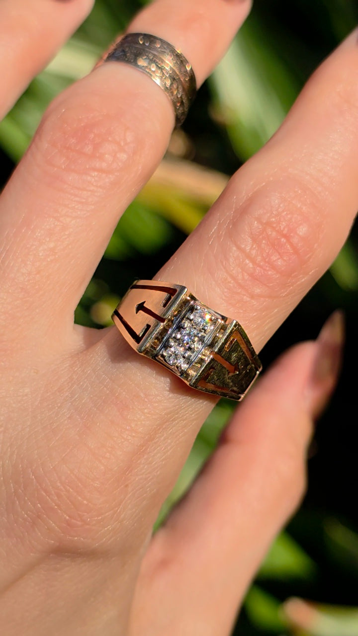 A close-up of a hand wearing a geometric gold ring with a central cluster of clear stones, set against a blurred green background.