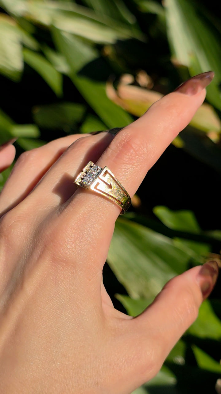 A gold ring with a clear gemstone set on a person's finger, photographed outdoors against a background of green leaves.
