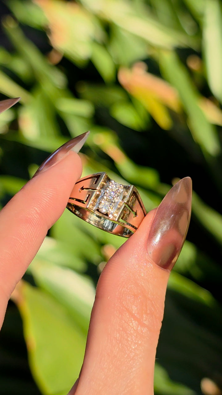 A close-up of a hand with long manicured nails holding a gold ring with a square-cut clear gemstone, set against a blurred background of green foliage.