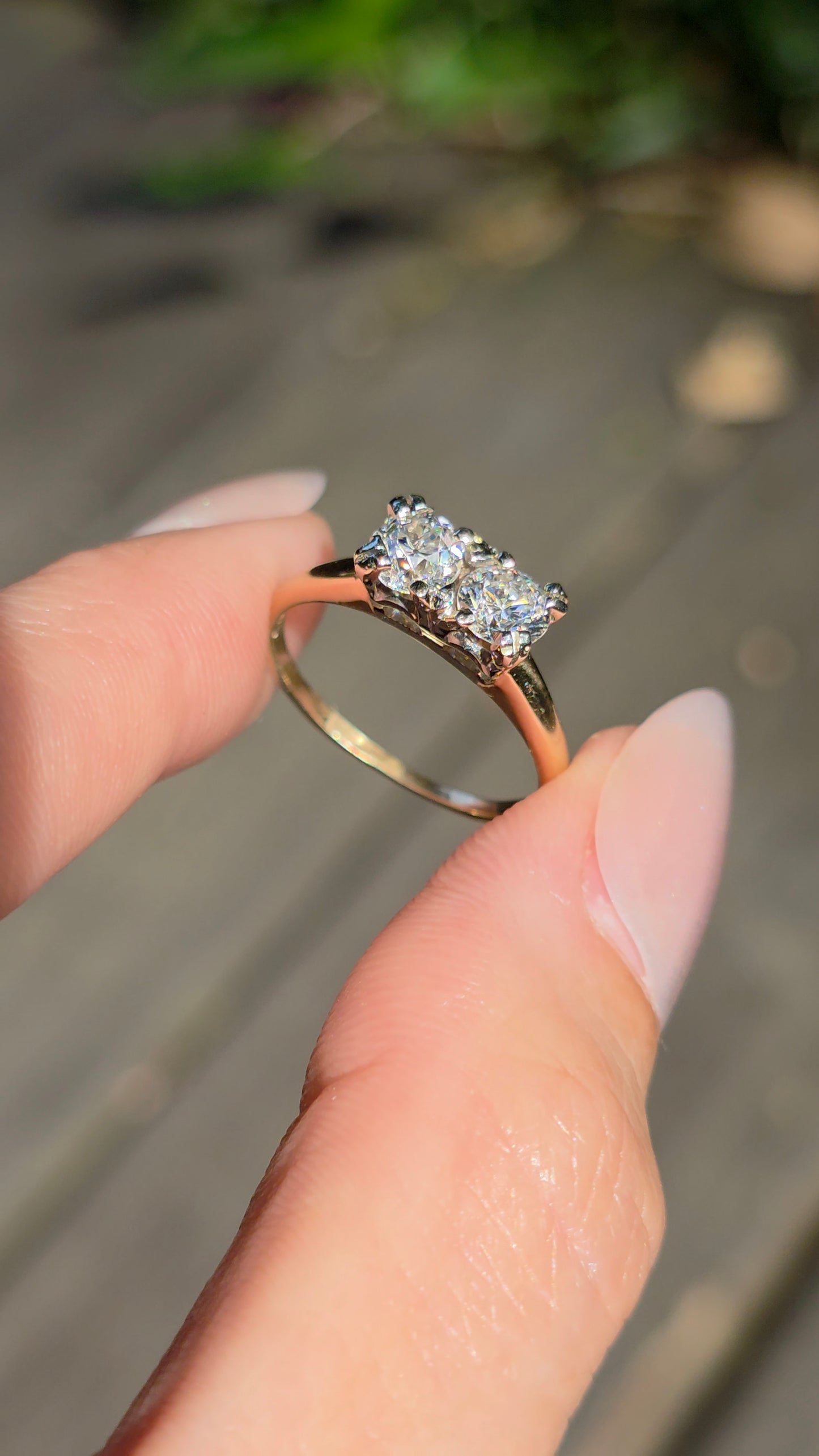 A close-up of a hand holding a rose gold ring with a three-stone diamond setting against a blurred outdoor background.
