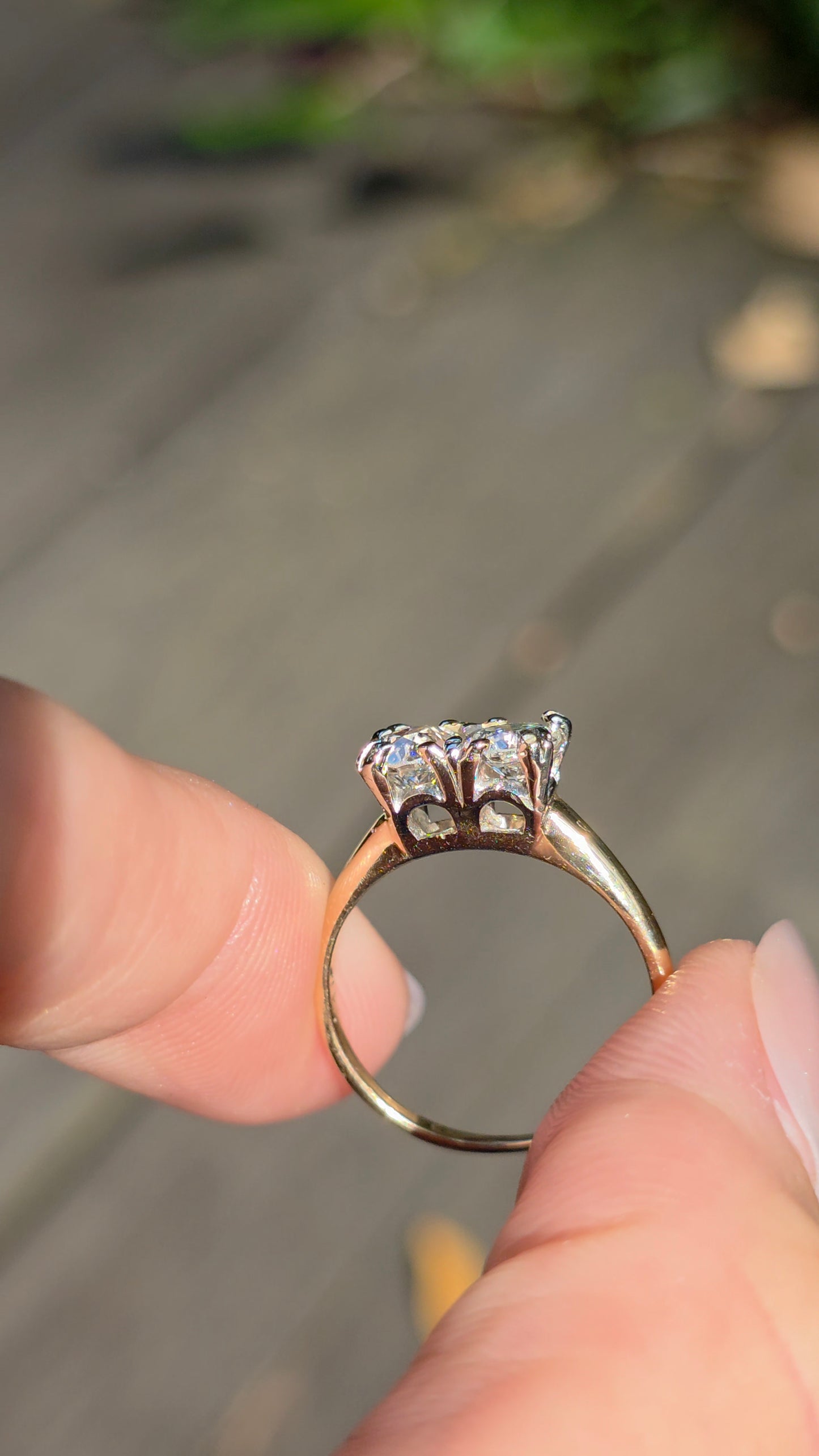 A close-up of a gold ring with a cluster of clear gemstones held between fingers against a blurred outdoor background.
