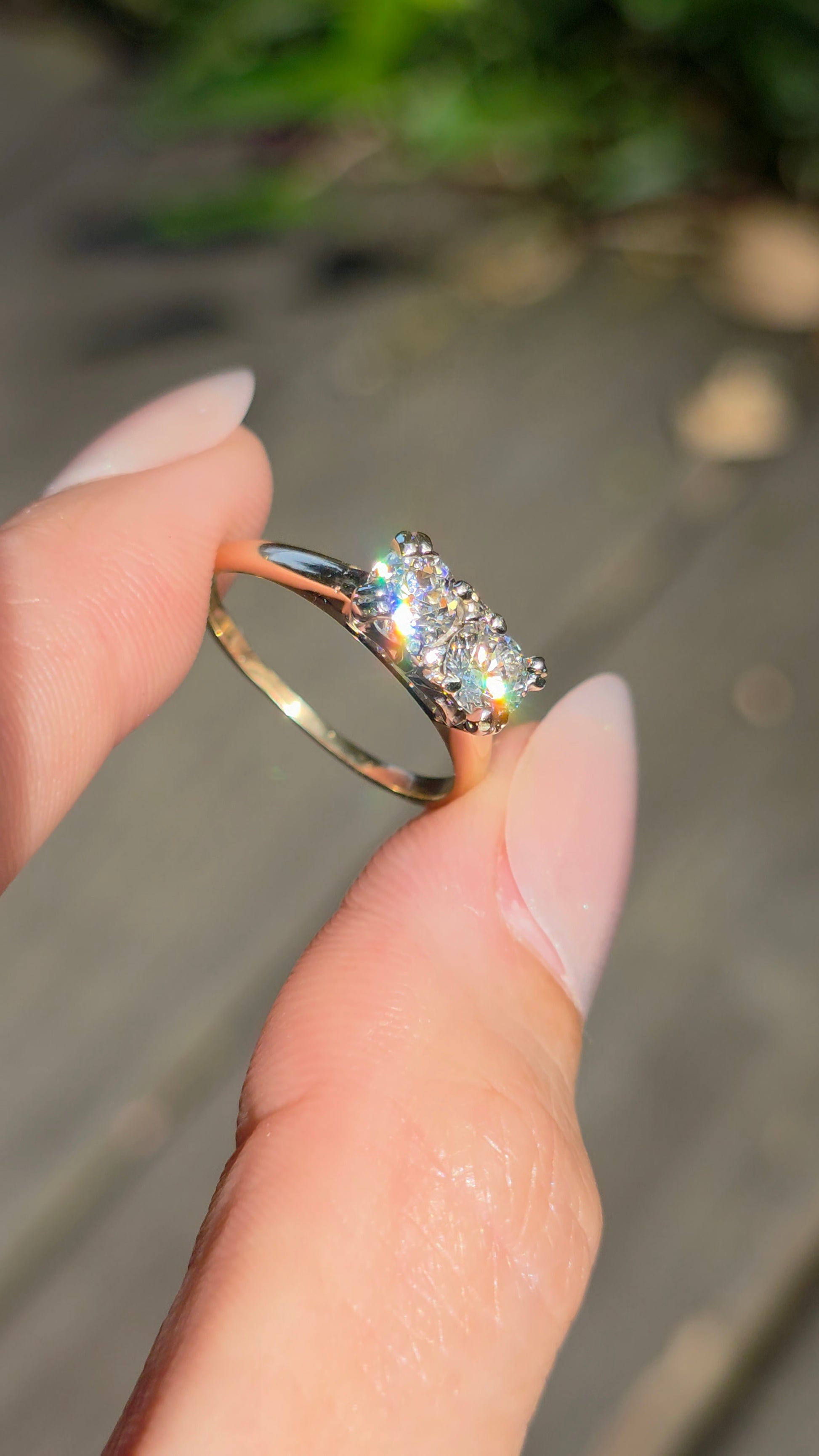 A close-up of a hand with manicured nails holding a rose gold ring with a sparkling cluster of clear gemstones, set against a blurred outdoor background.