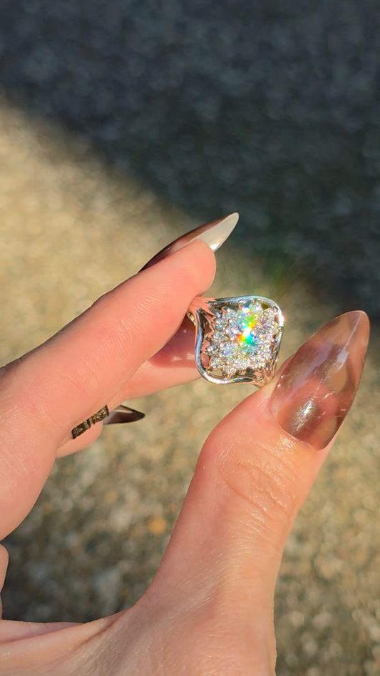 A close-up of a hand with long manicured nails holding a silver ring featuring a square cushion-cut clear gemstone with a colorful iridescent center, set against a blurred outdoor background.