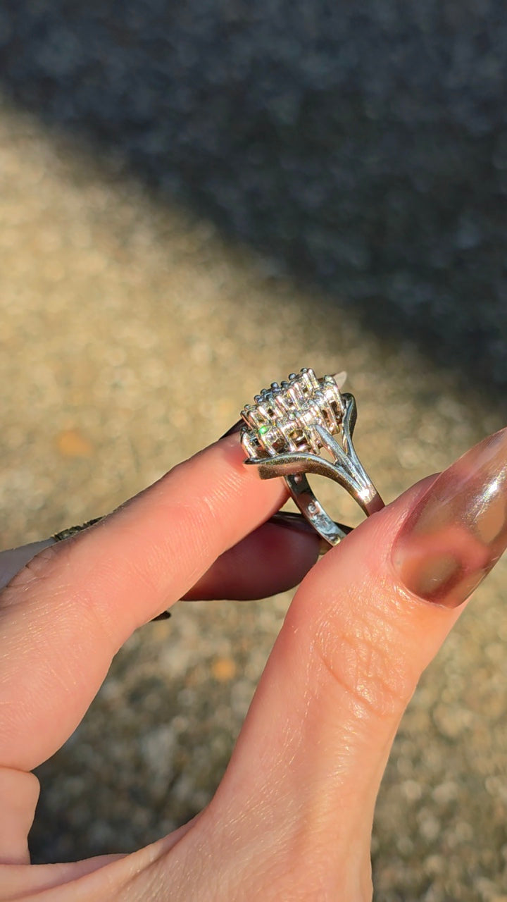 A close-up of a hand holding a silver-toned ring with a cluster of clear gemstones, photographed outdoors against a blurred ground background.