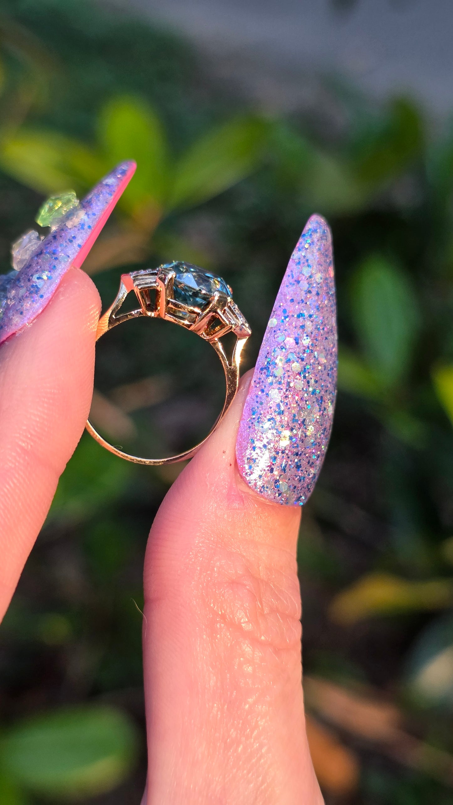 Hand with glittery nail polish holding a gold ring with a blue gemstone against a blurred green background