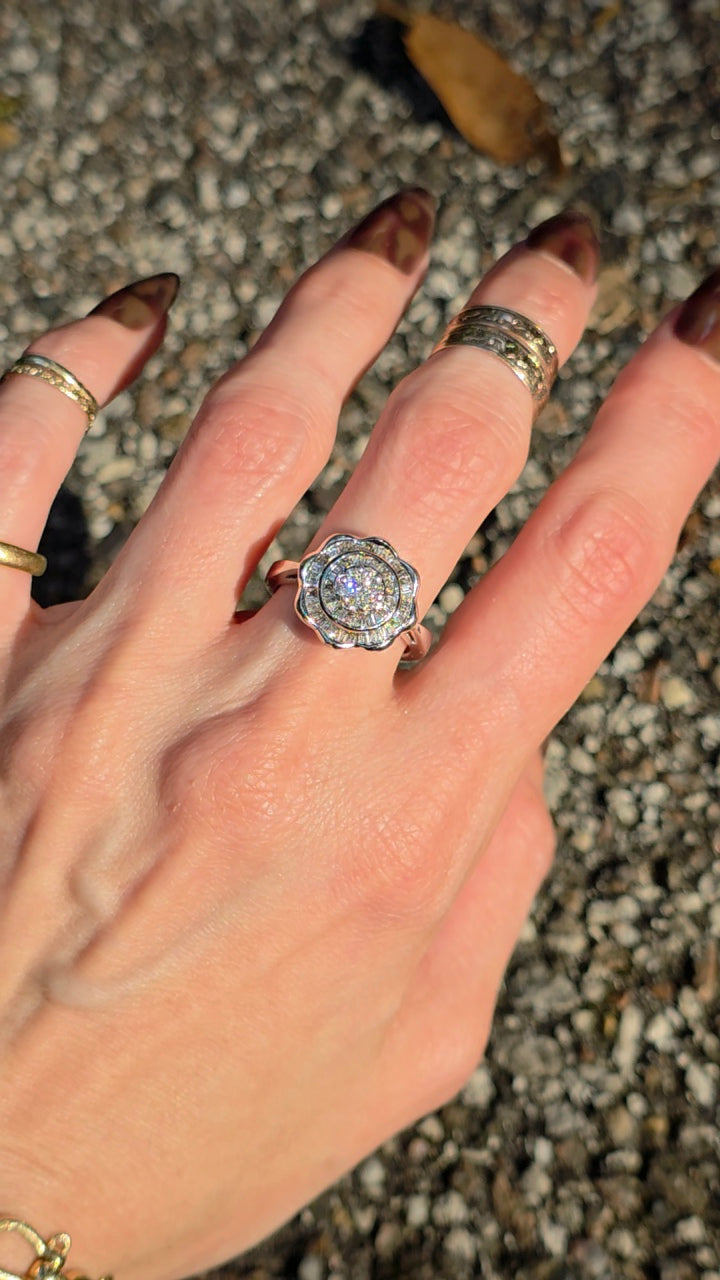 A close-up of a hand wearing a silver-toned flower-shaped ring with a clear center stone, along with other rings, set against a gravel background.