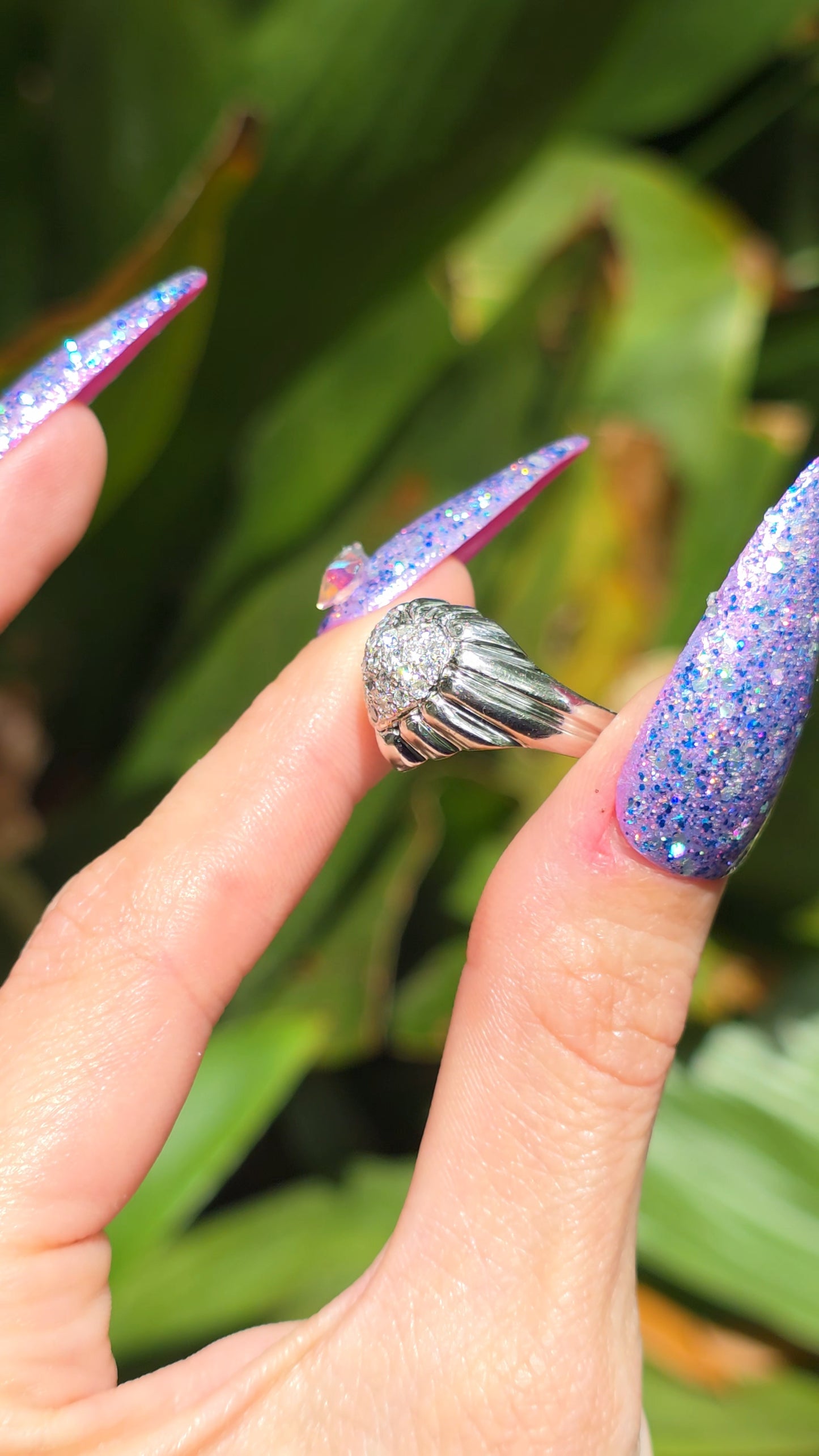 Gold heart ring with a large gemstone on a finger with purple glittery nail polish against a green leafy background