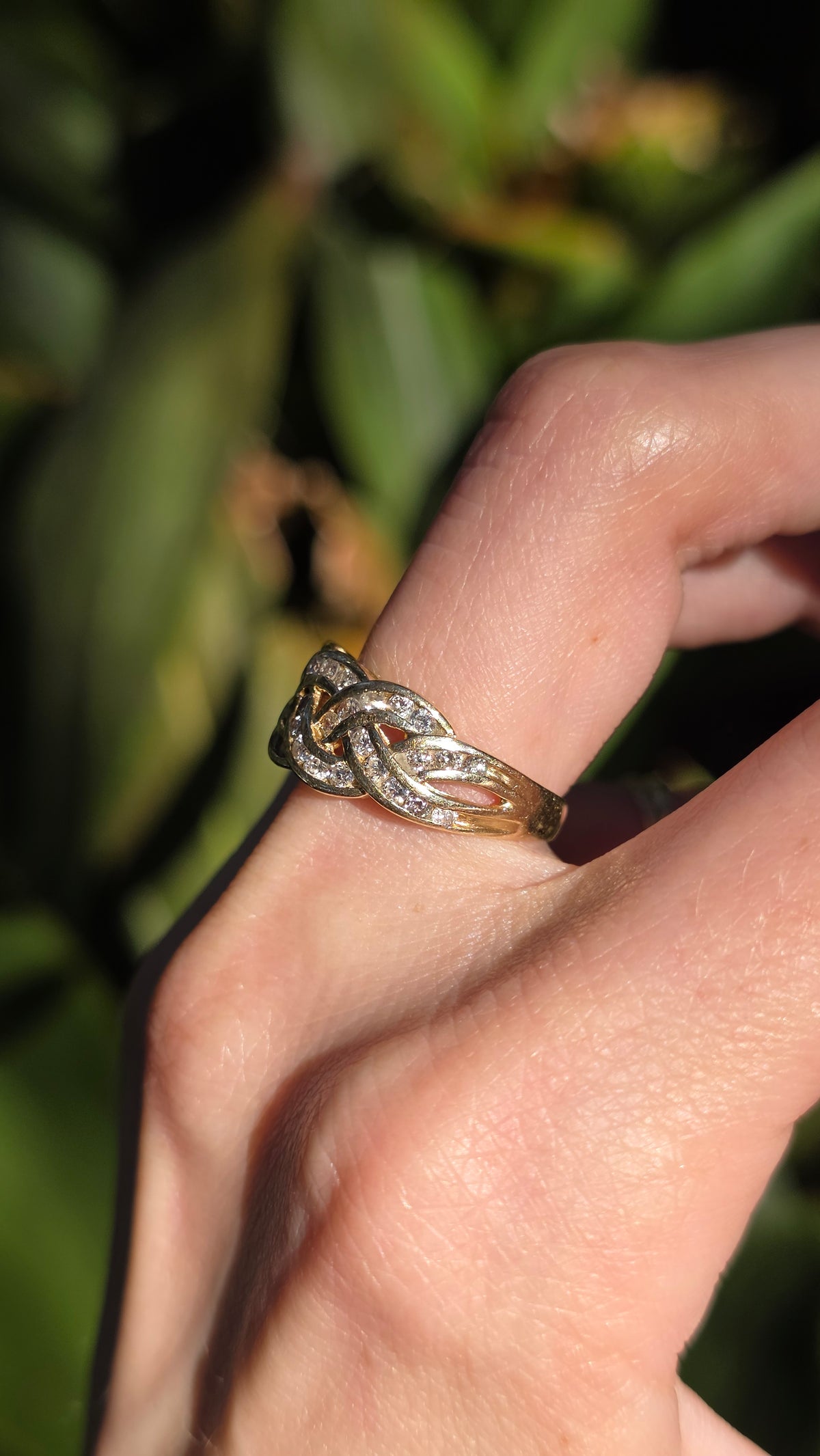 A close-up of a hand wearing a gold and silver interwoven knot ring, set against a blurred green outdoor background.
