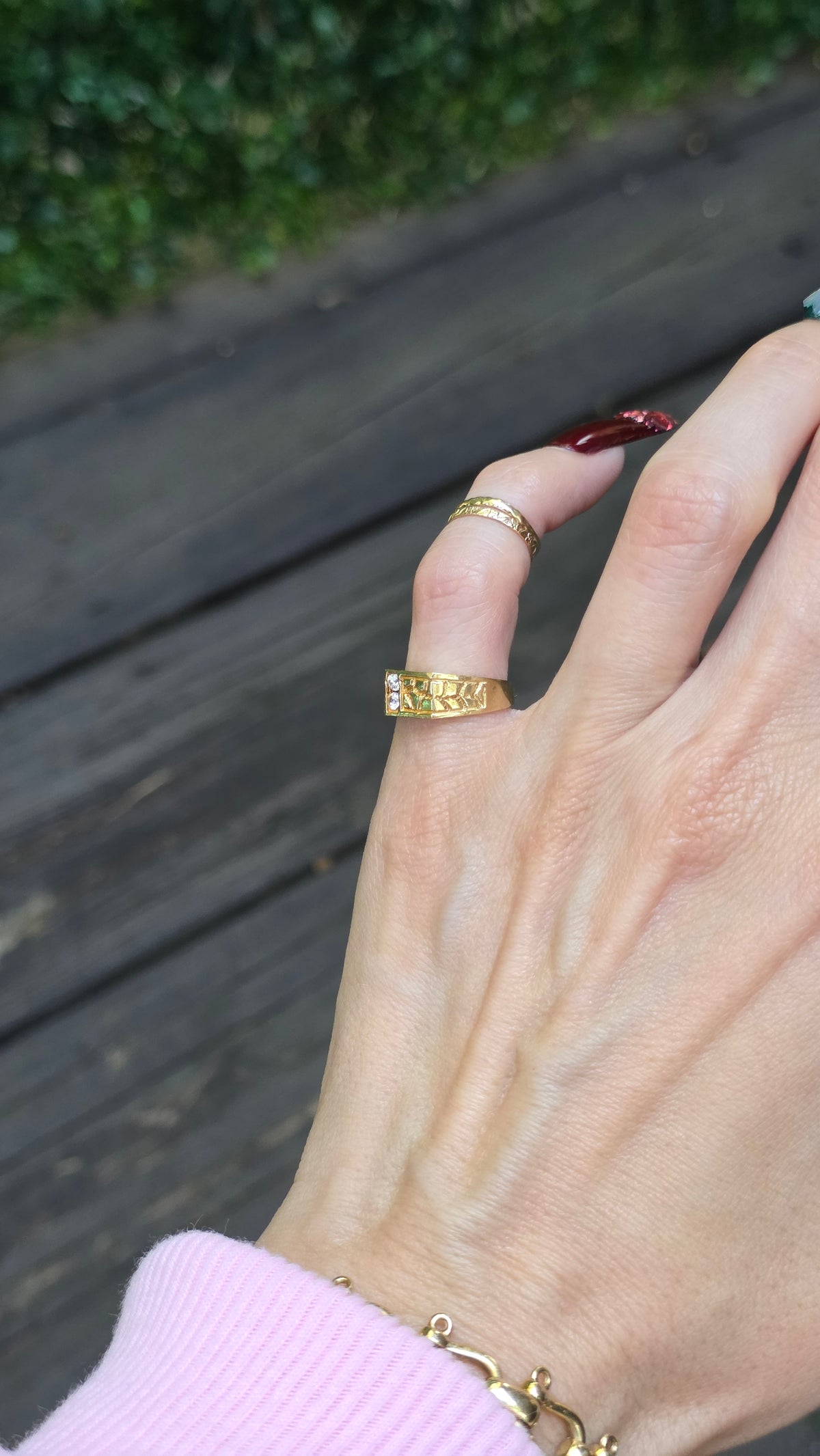 A close-up of a person's hand wearing two gold rings, one of which has a textured or engraved band, shown outdoors on a wooden deck.