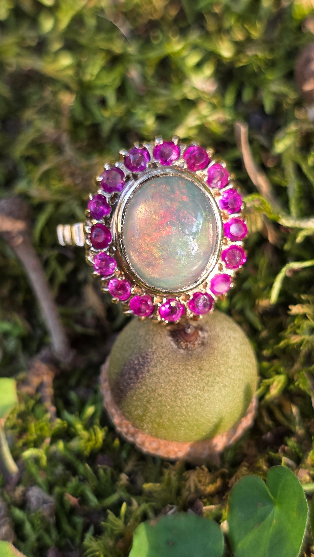 A gold ring with a large iridescent opal center stone and a halo of pinkish-purple gemstones, resting on green moss outdoors.