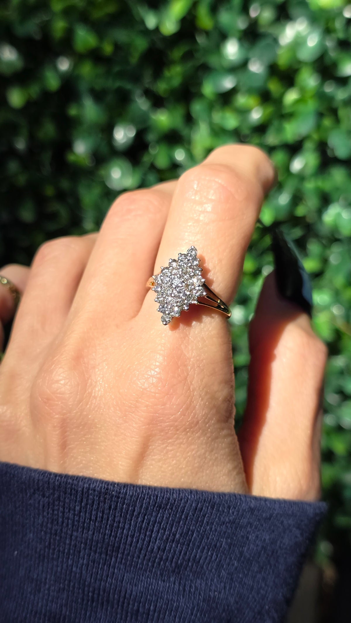 A close-up of a hand wearing a sparkling diamond cluster ring with a gold band, set against a blurred green hedge background.