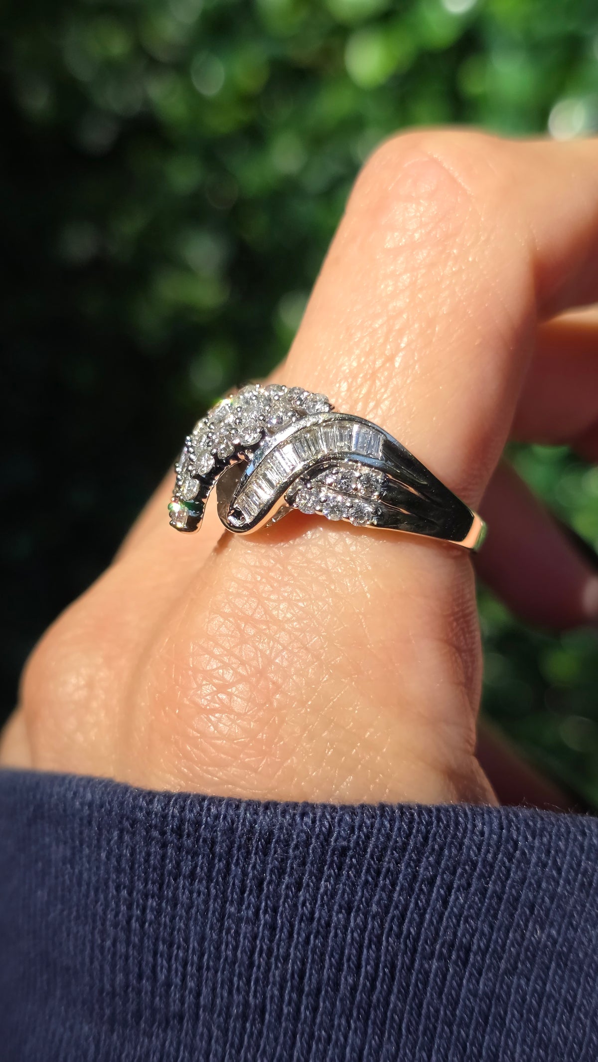 A close-up of a hand wearing a silver-toned ring with a curved, diamond-encrusted design, set against a blurred green outdoor background.