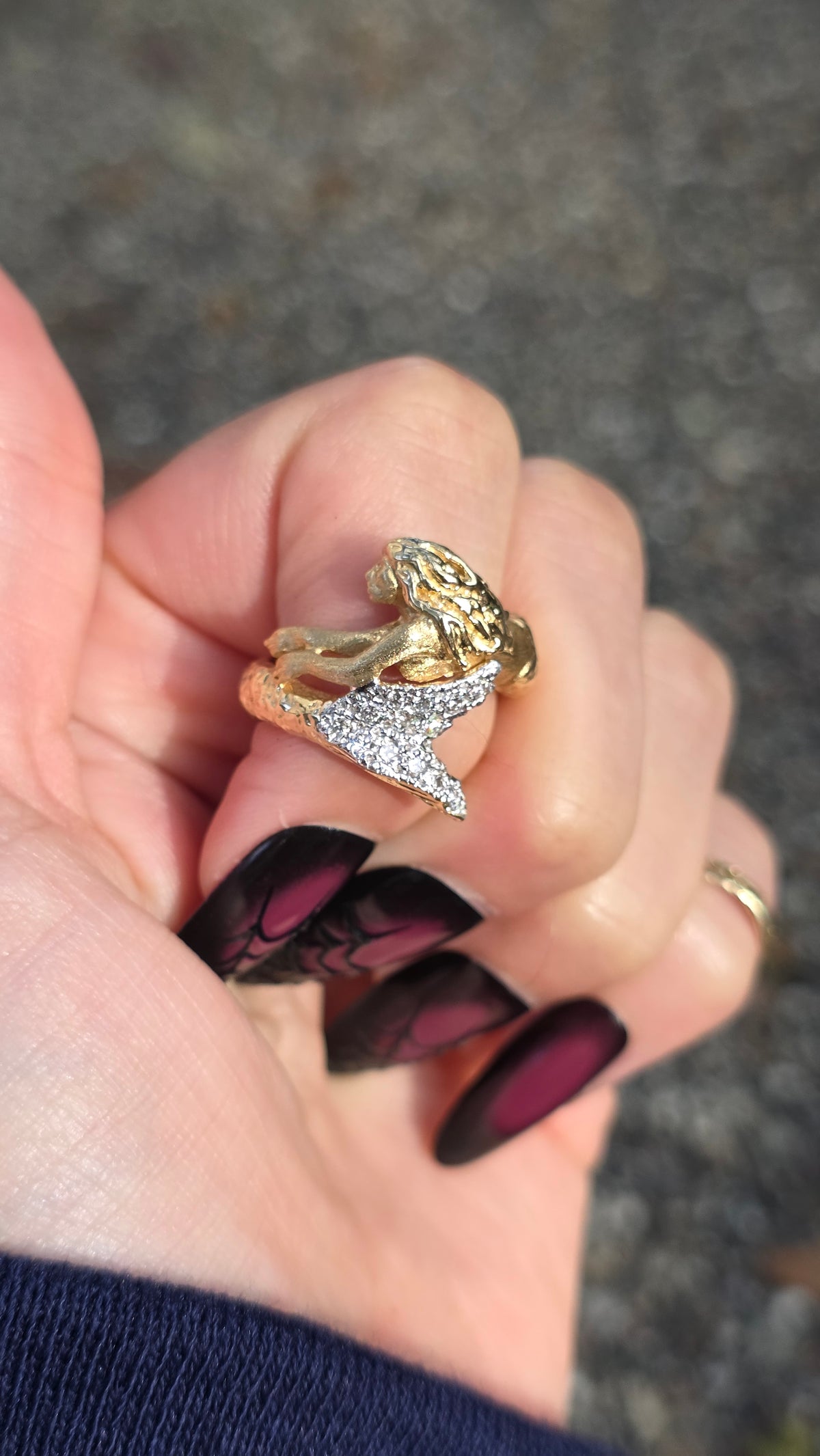 A gold and silver ring featuring a mermaid design, held in a hand with dark patterned nails against a blurred gray asphalt background.