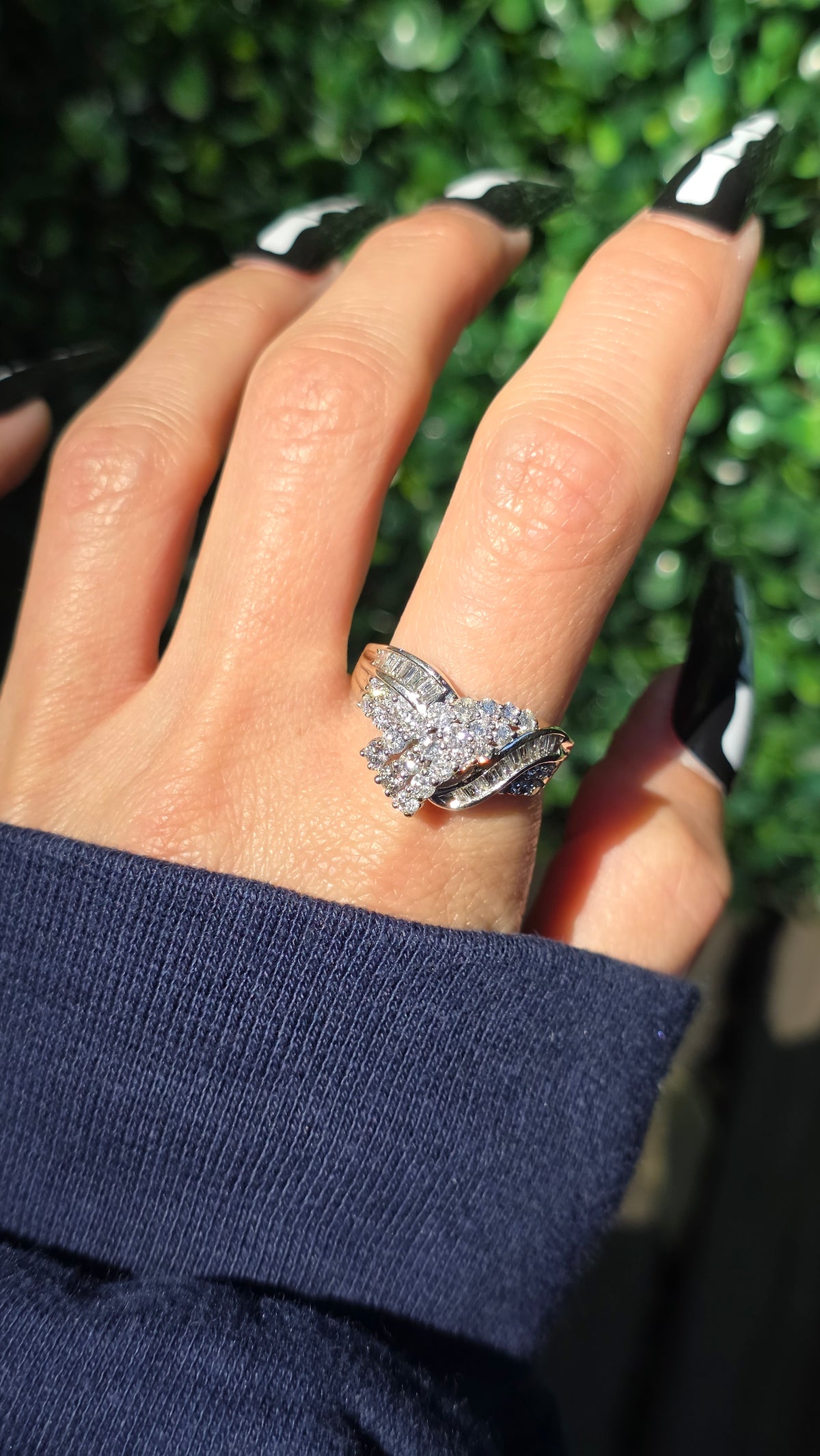A silver-toned ring with a wing-like or butterfly-inspired design set with clear stones, worn on a hand with black-and-white patterned nail polish against a background of green foliage.