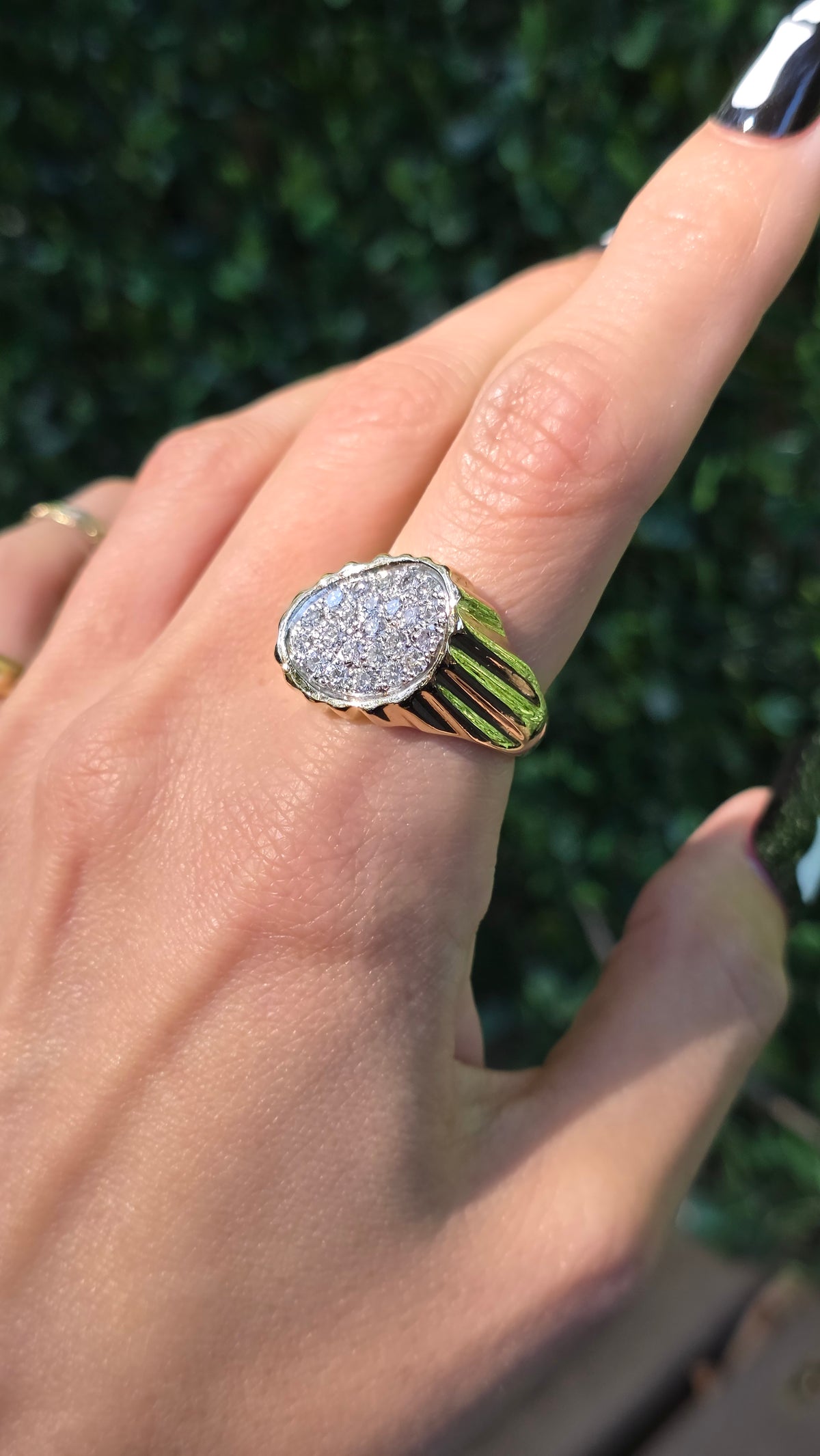 A close-up of a hand wearing a gold signet ring with a pavé diamond oval face, set against a blurred green hedge background.