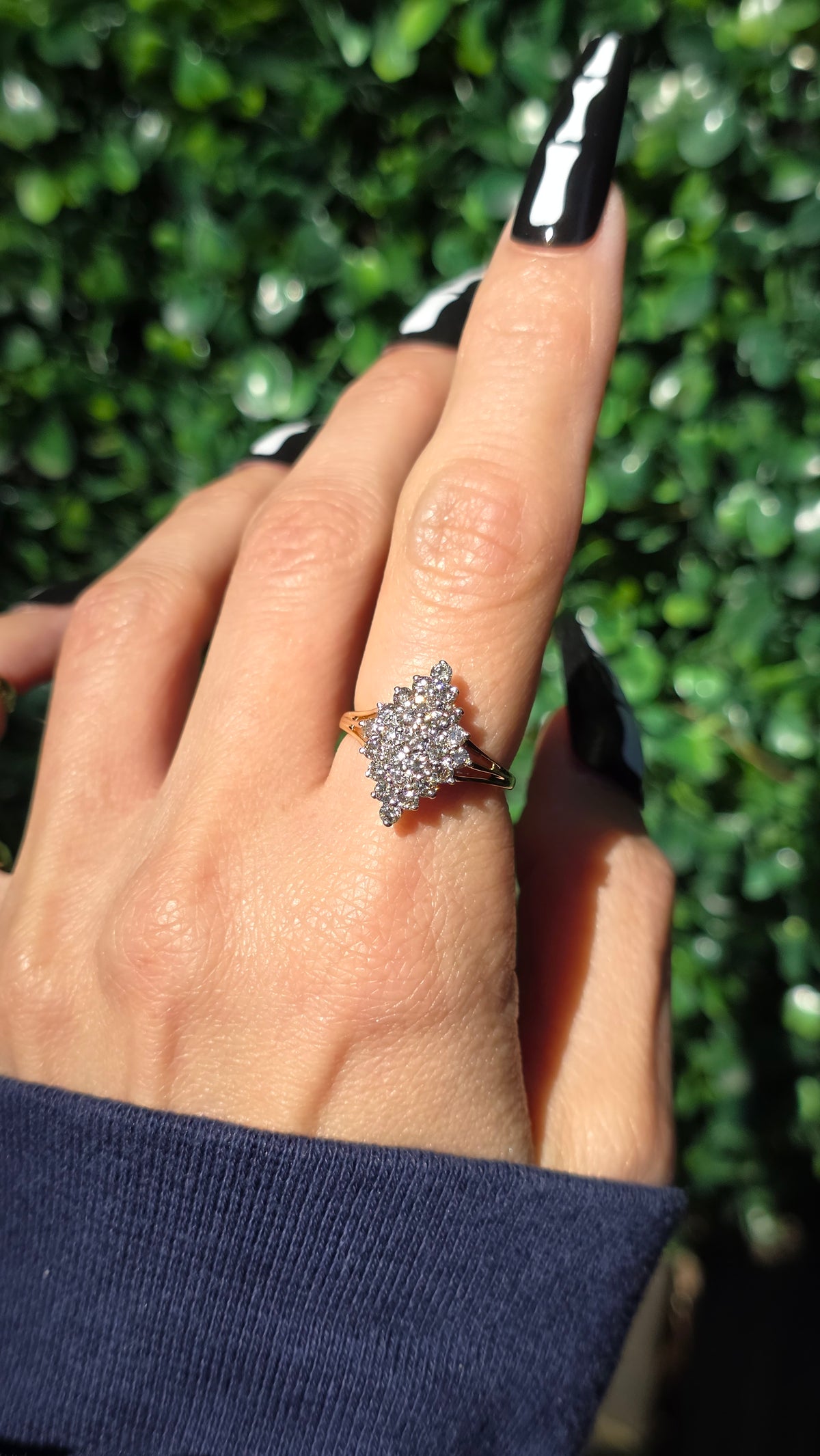 A close-up of a hand wearing a gold ring with a pavé diamond star-shaped cluster, set against a background of green foliage.