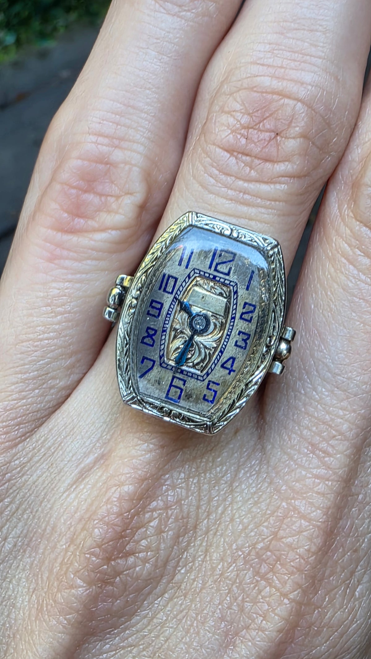 A vintage-style ring designed to look like a watch face, worn on a person's finger. The ring has a silver-toned textured frame, a light blue dial with blue Arabic numerals, and a central skeletonized watch movement.