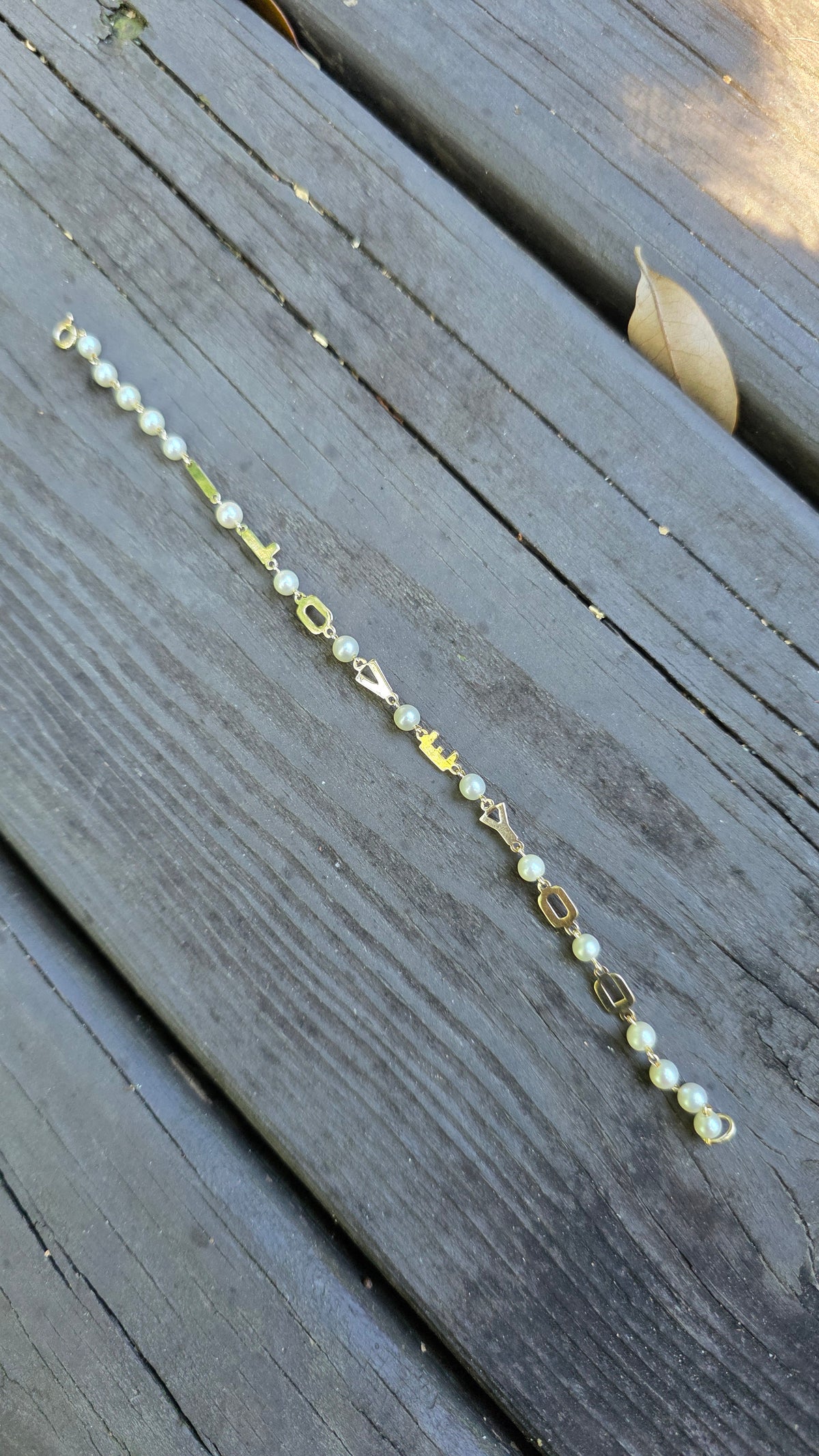 A delicate gold chain bracelet with small white pearl-like beads and clear faceted beads, laid diagonally on weathered gray wooden planks.