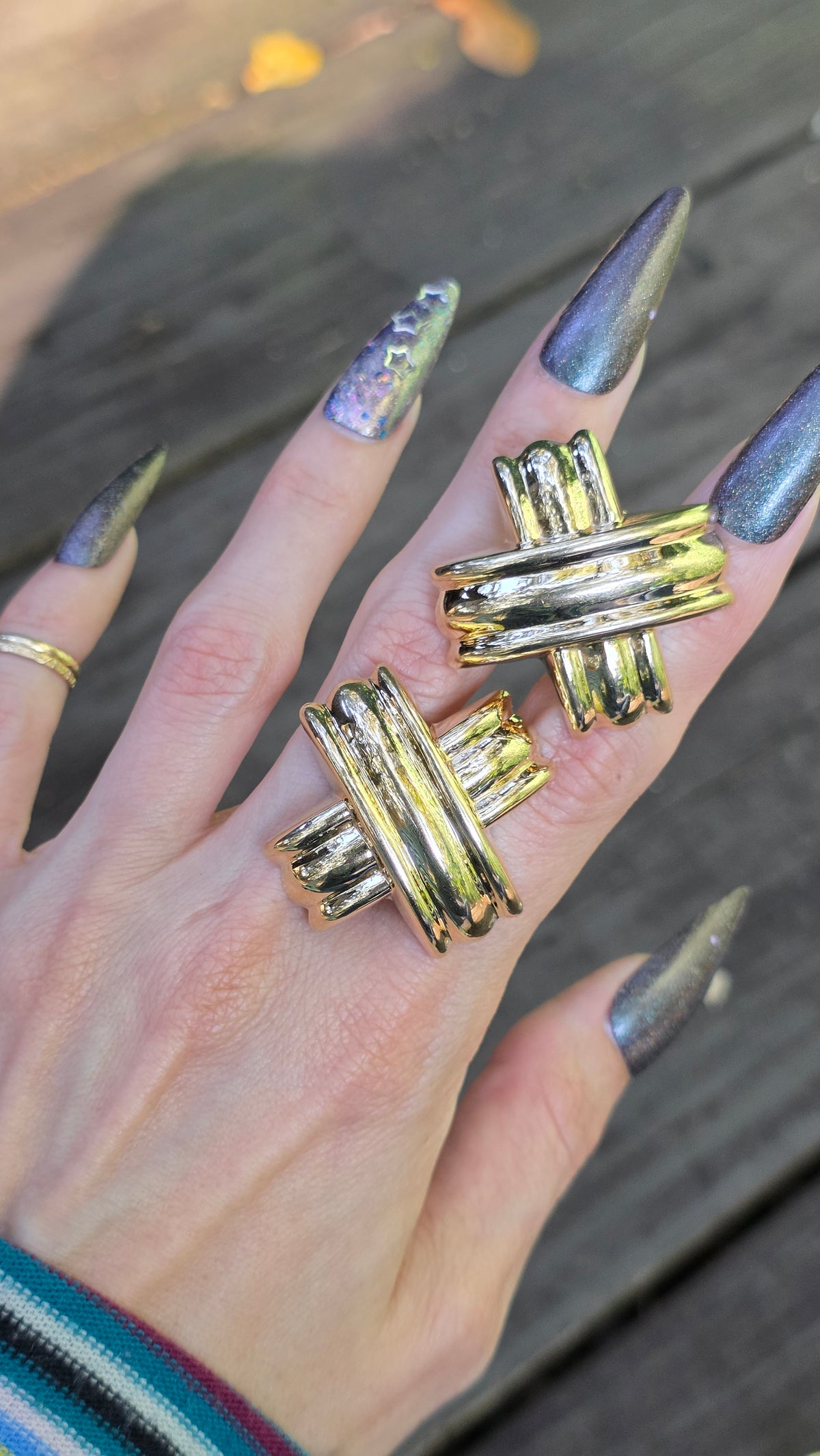 A close-up of a hand wearing two chunky gold earrings with a twisted, knotted design, set against a blurred wooden background.