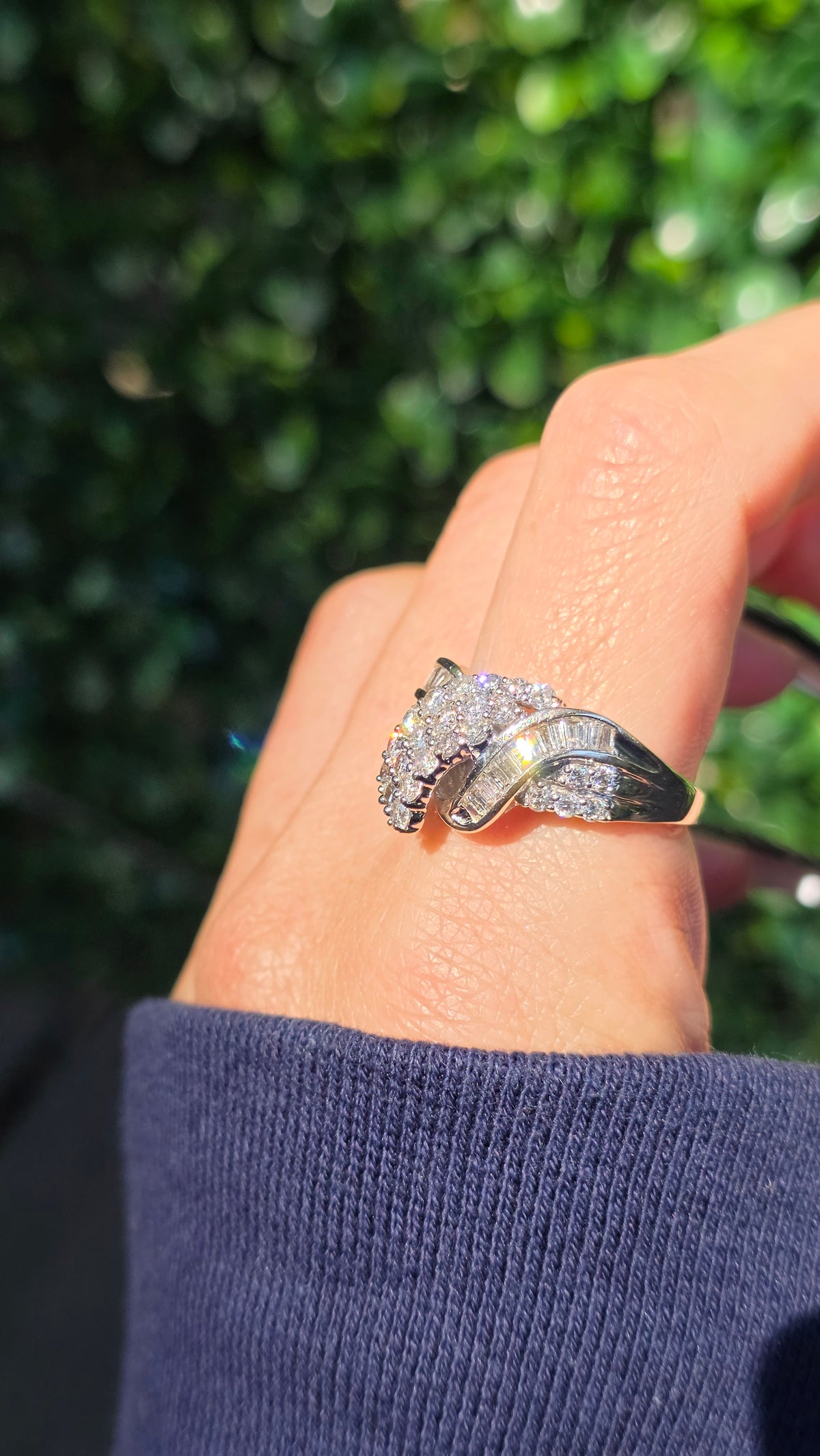 A silver-toned ring with a pavé diamond cluster setting worn on a person's hand, with a blurred green outdoor background.