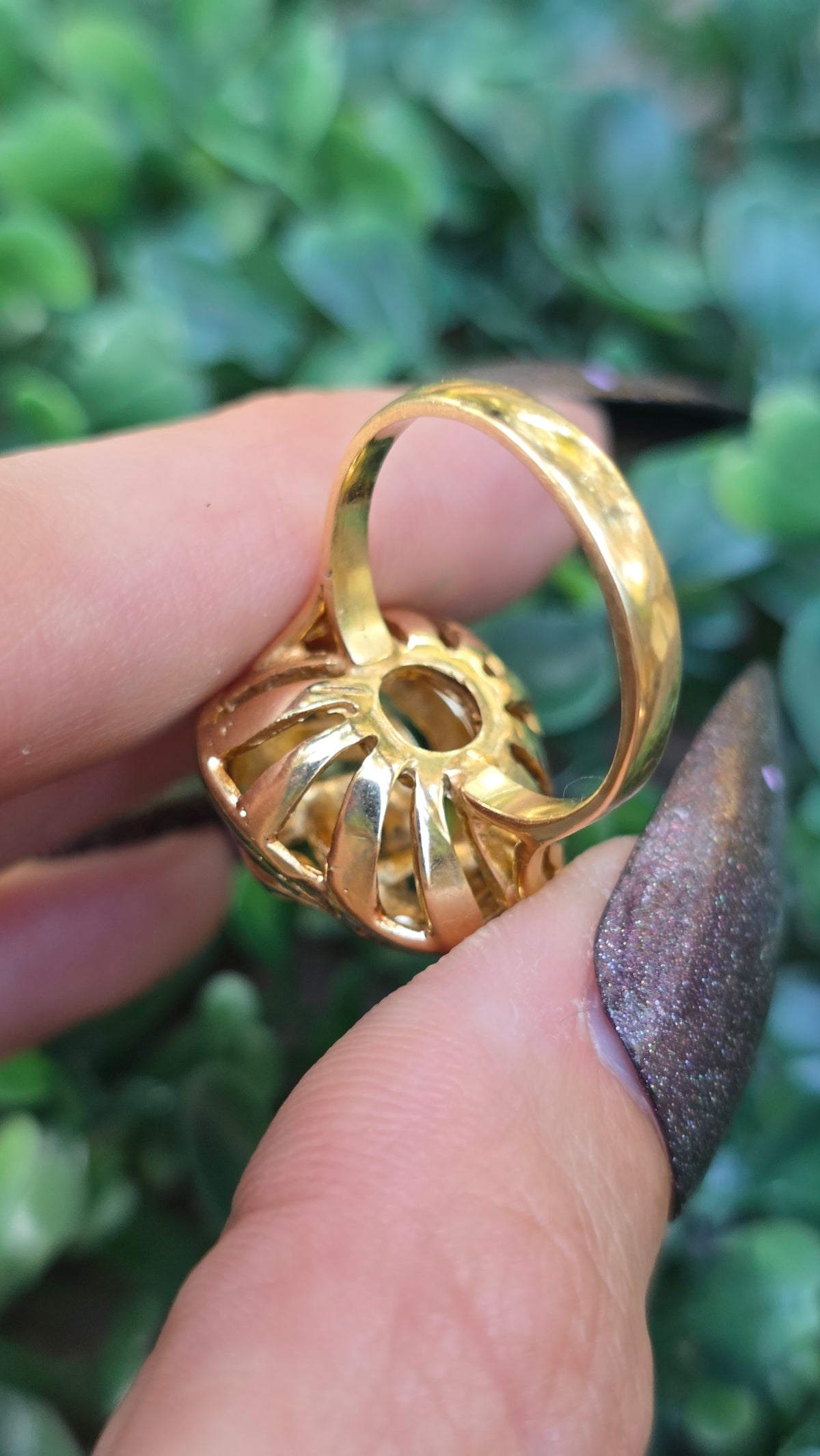 A close-up of a gold ring with a textured, ribbed pavé-style band, held between fingers against a blurred green leaf background.