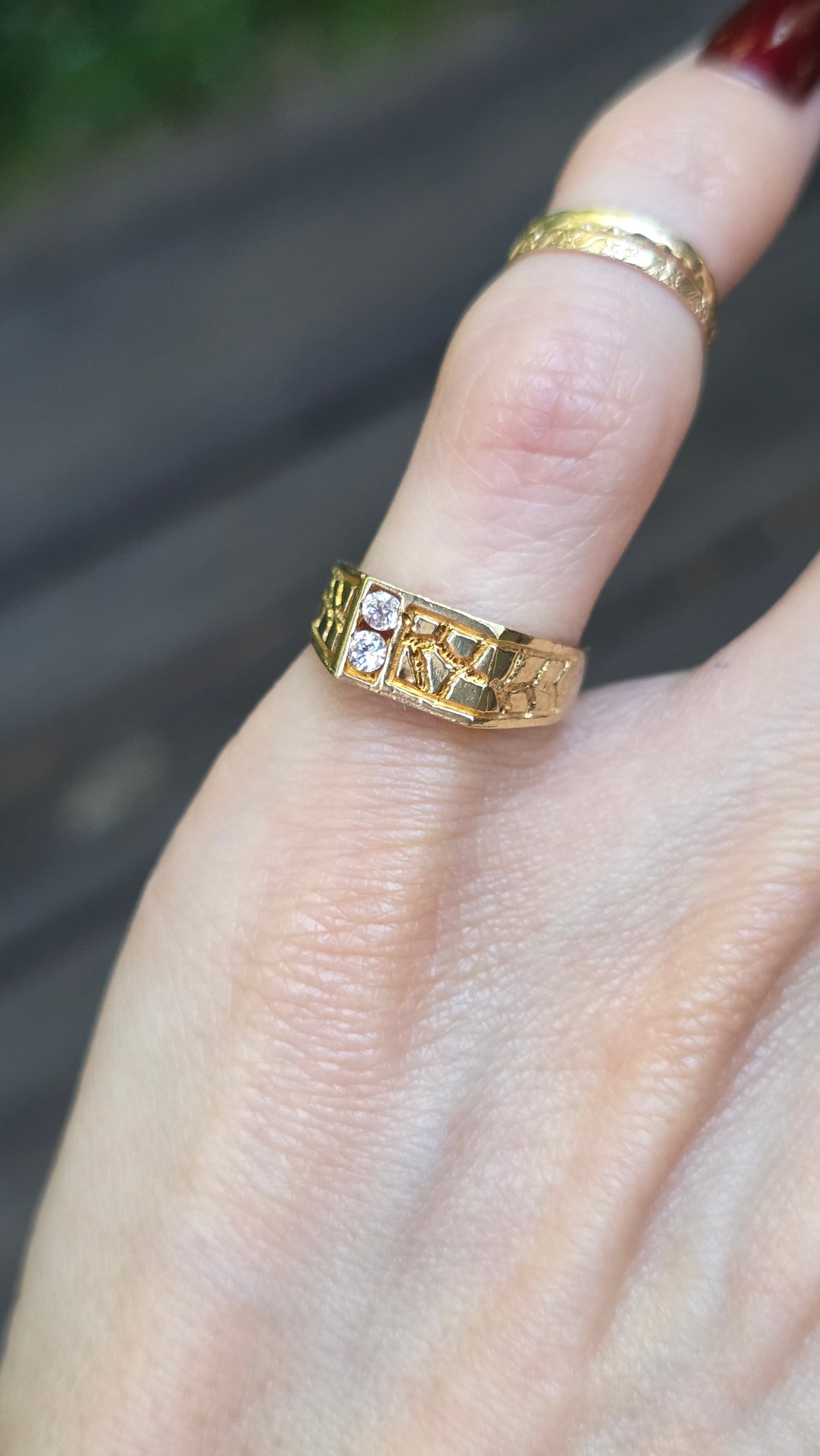 A close-up of a hand wearing a textured yellow gold ring with small clear stones, with a second gold band on the middle finger, against a blurred outdoor background.