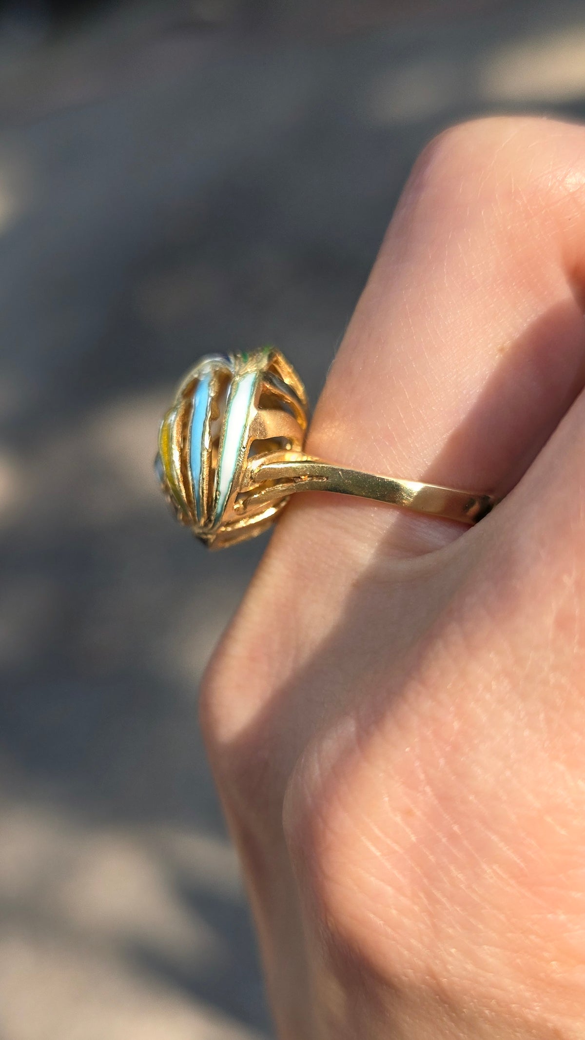 A gold ring with a textured, basket-like openwork top and a light blue inlay, shown worn on a person's finger against a blurred outdoor background.