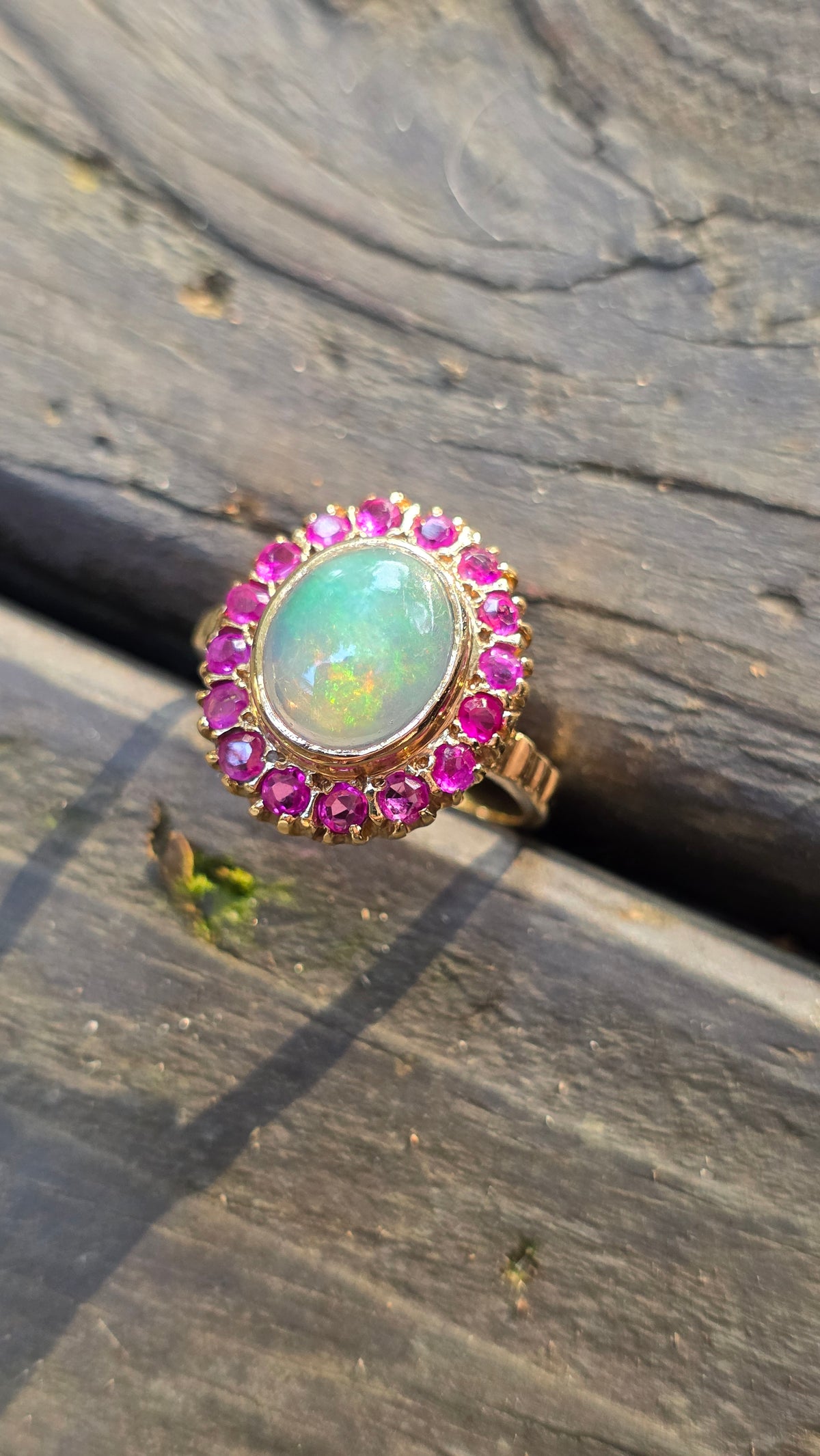 A gold ring featuring a large oval green opal center stone surrounded by a halo of pink gemstones, resting on a weathered dark wood surface.