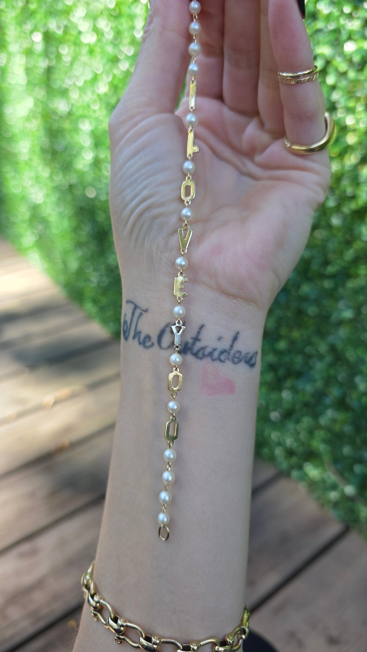 A gold chain bracelet with small white pearls and decorative charms, shown worn on a person's wrist against a blurred outdoor background of green foliage and wooden decking.