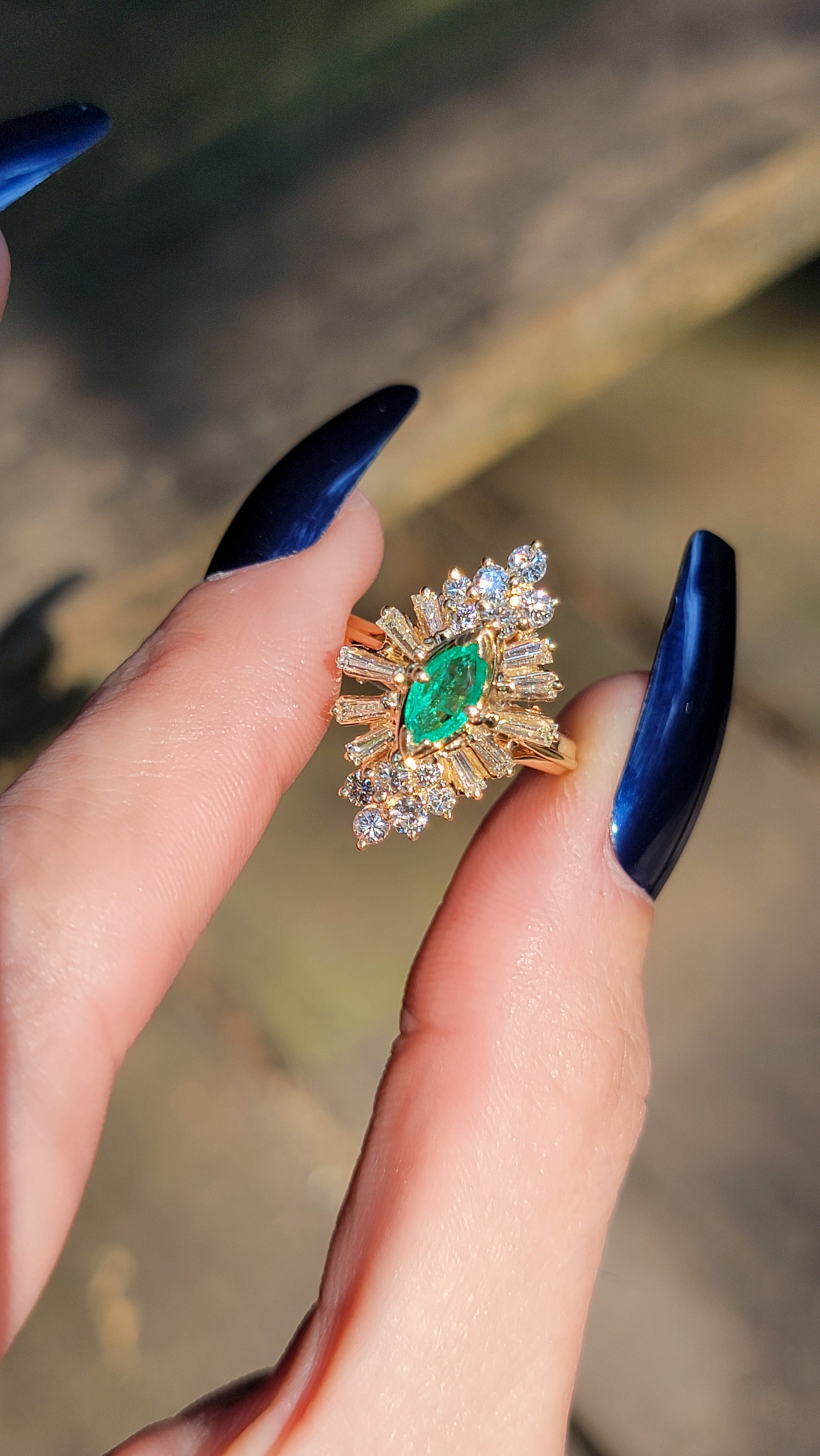 Gold ballerina ring with emerald stone held between fingers against a blurred background
