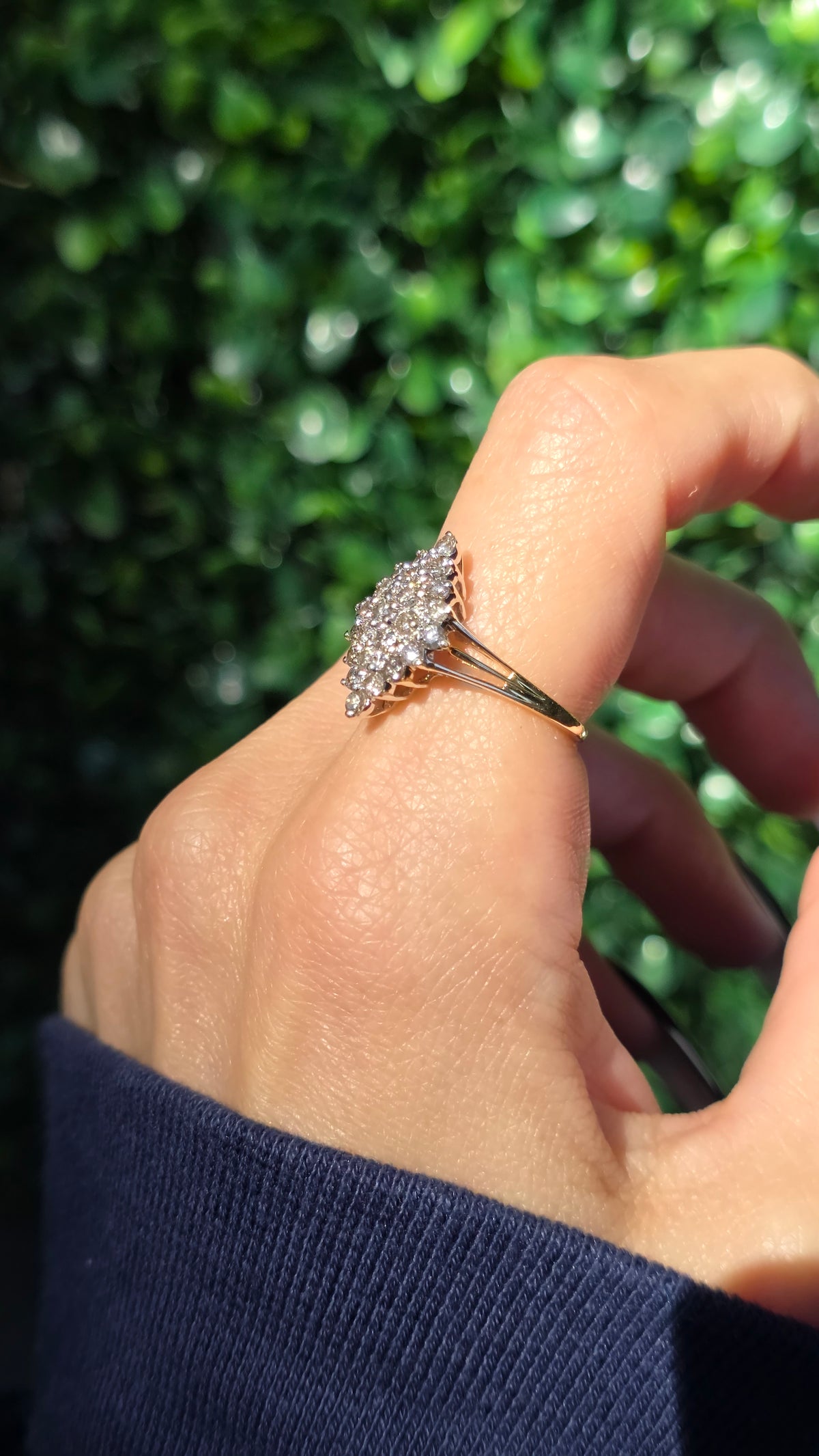 A close-up of a hand wearing a gold ring with a cluster of clear gemstones, set against a blurred green hedge background.
