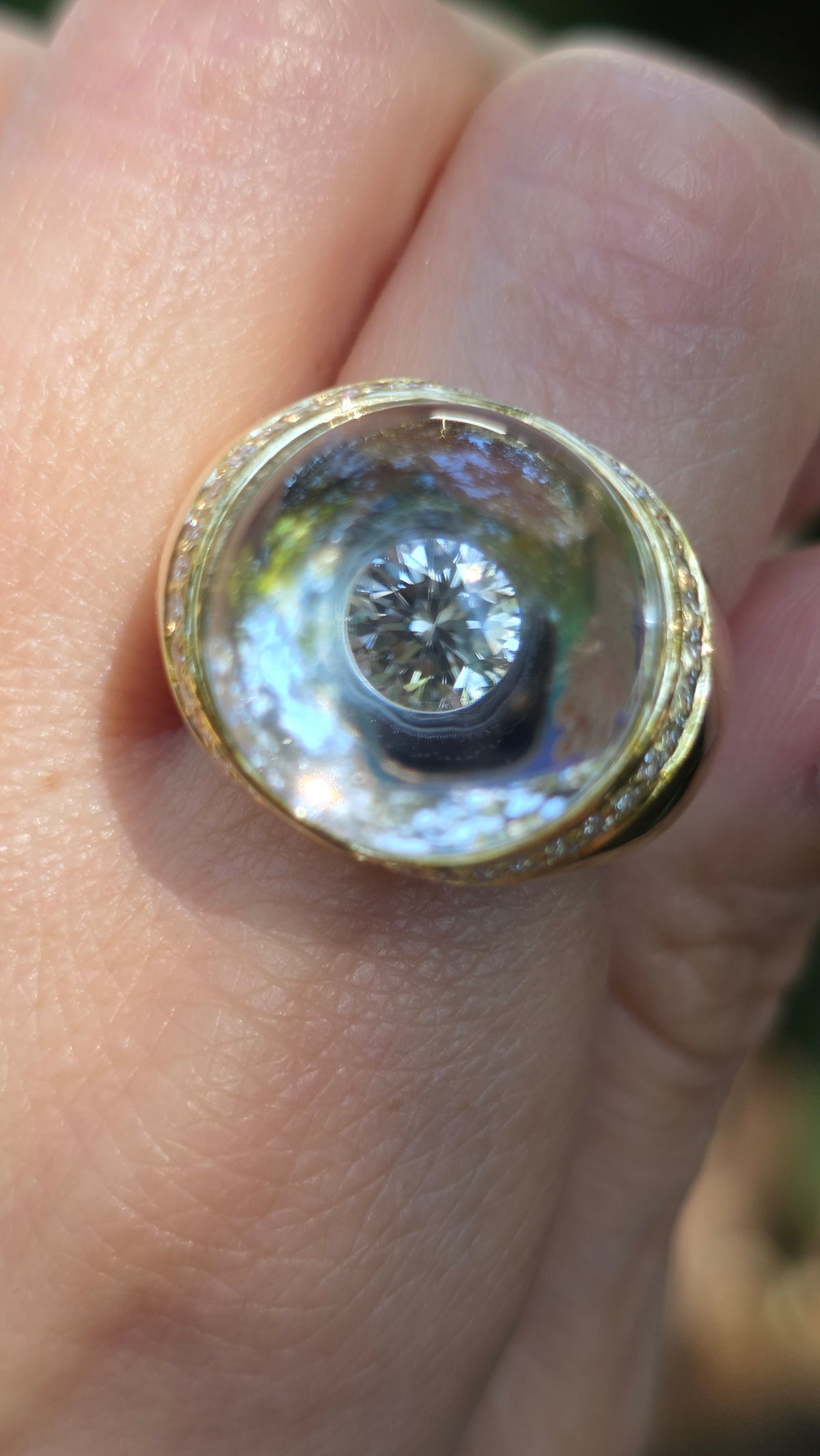 A close-up shot of a gold ring with a clear, dome-shaped iridescent inlay and a central clear gemstone, worn on a finger.
