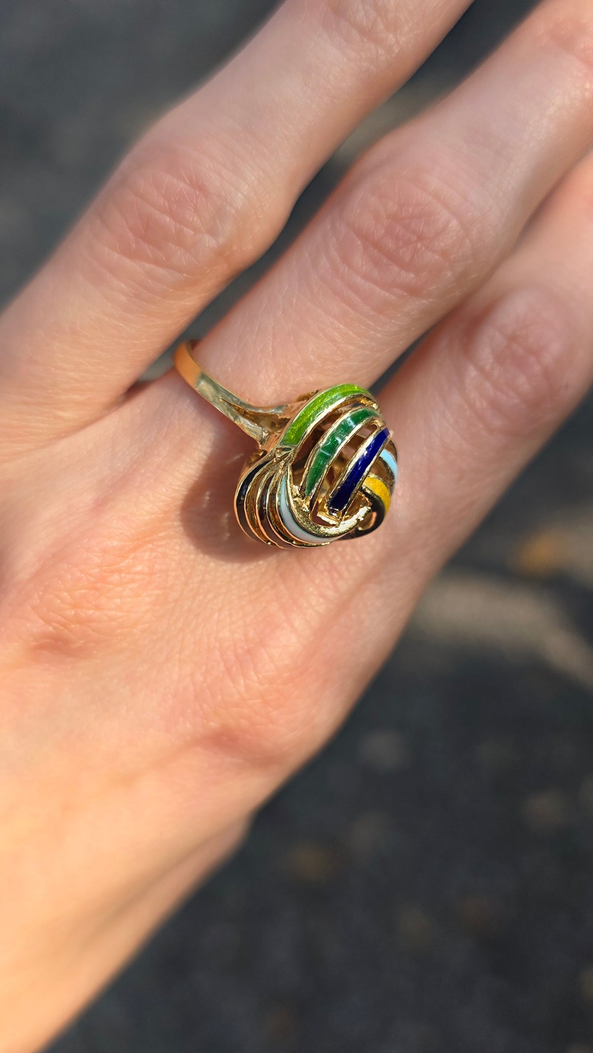 A close-up of a hand wearing a gold ring with a heart-shaped, multi-colored enamel design featuring green, blue, yellow, and white swirls.