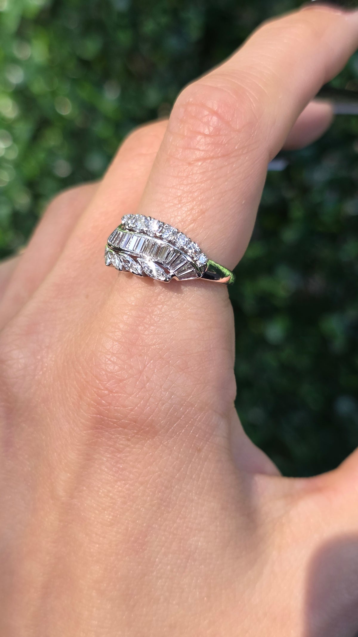 A close-up of a hand wearing a silver-toned ring with baguette-cut clear stones, set against a blurred green outdoor background.