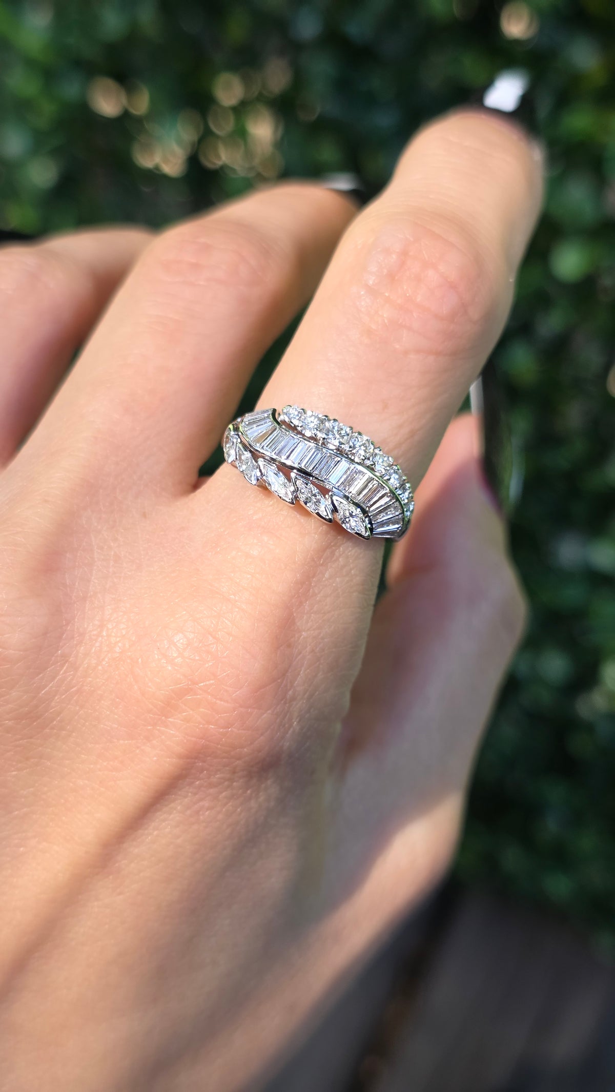 A close-up of a hand wearing a silver-toned diamond ring with baguette-cut and round diamonds, set against a blurred green outdoor background.