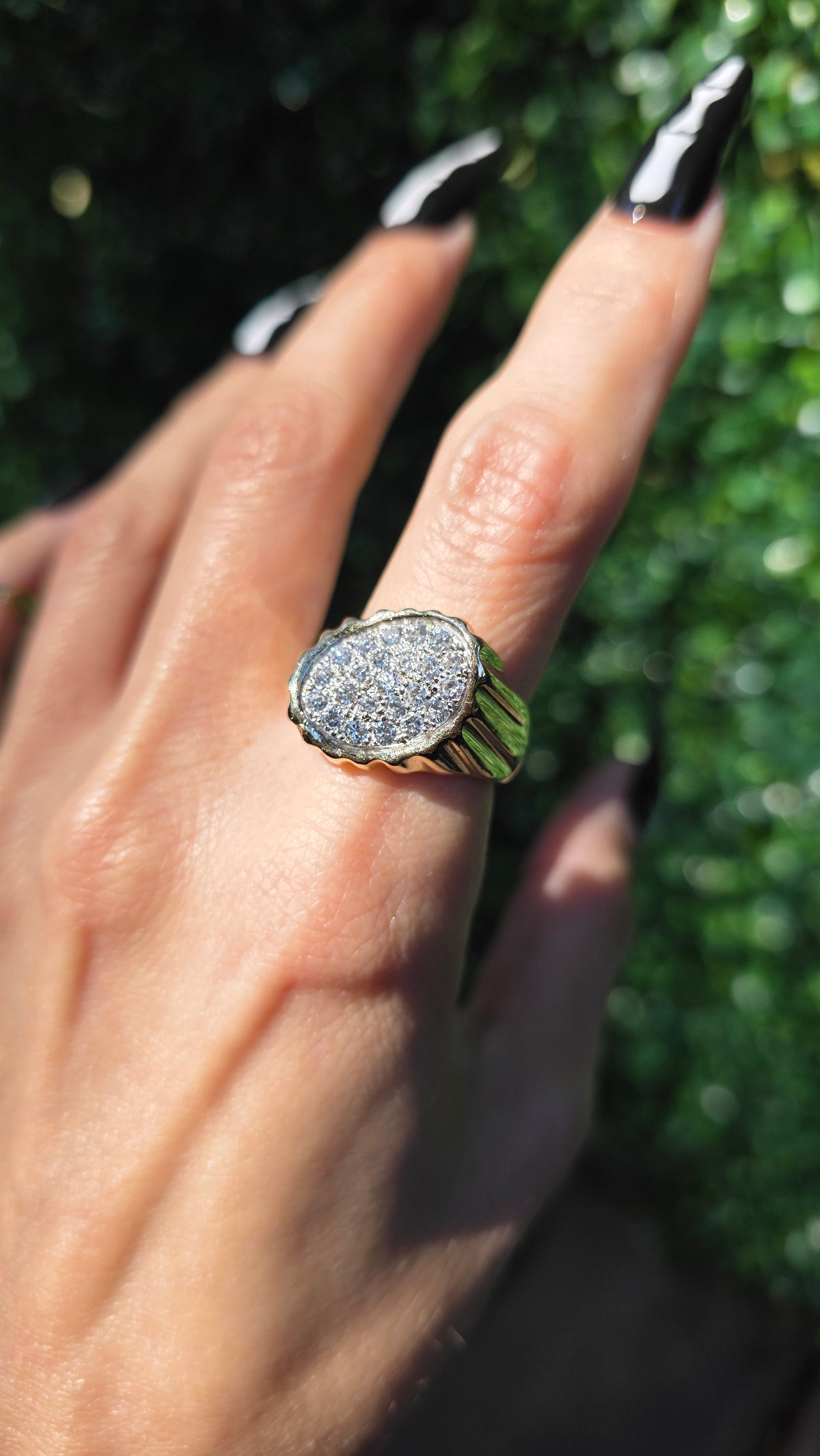 A close-up of a hand wearing a gold signet-style ring with a textured, diamond-like silver oval face, set against a blurred green outdoor background.