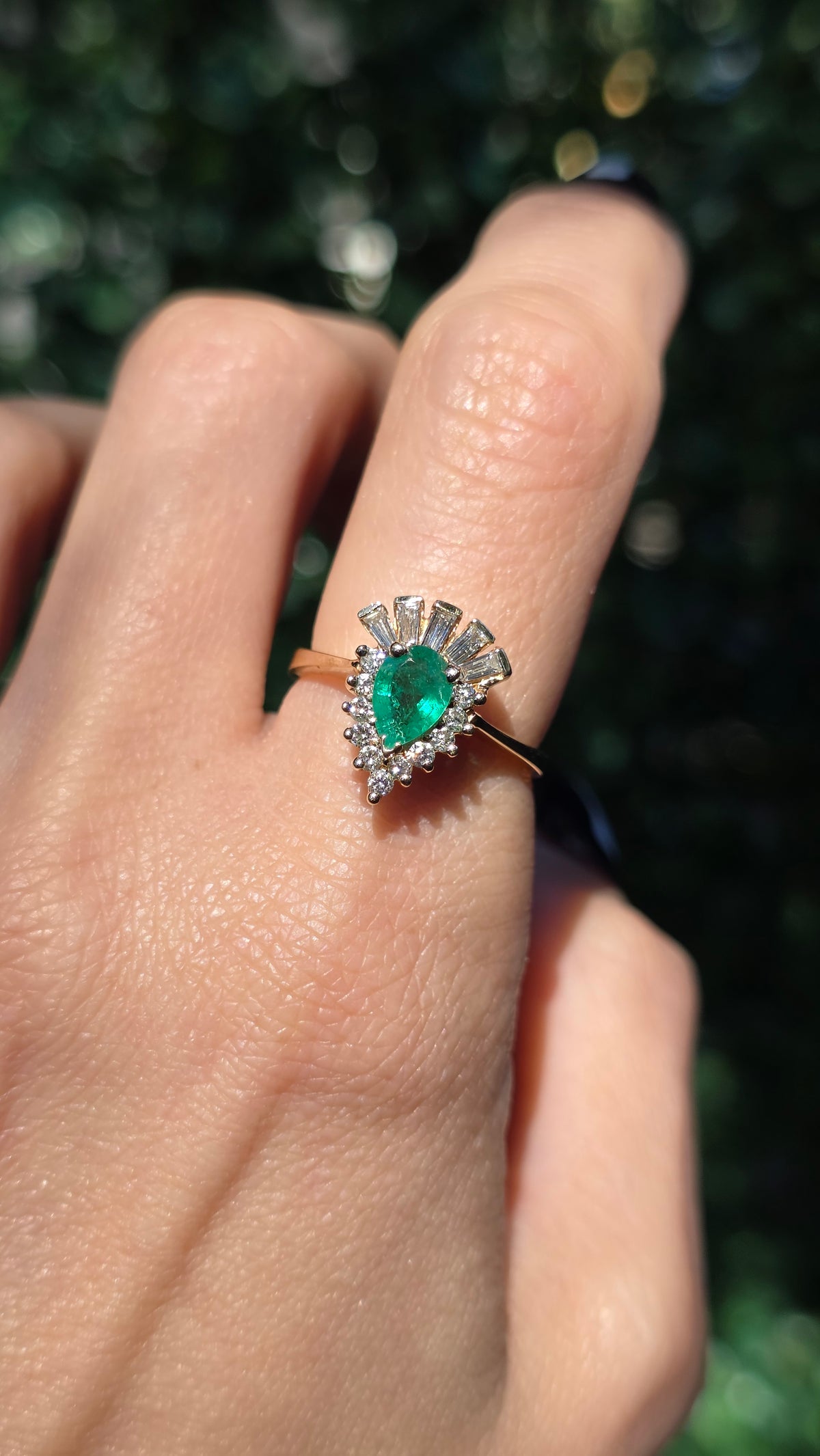 A close-up of a hand wearing a ring with a green teardrop-shaped gemstone and silver-toned metal settings, photographed outdoors against a blurred green background.