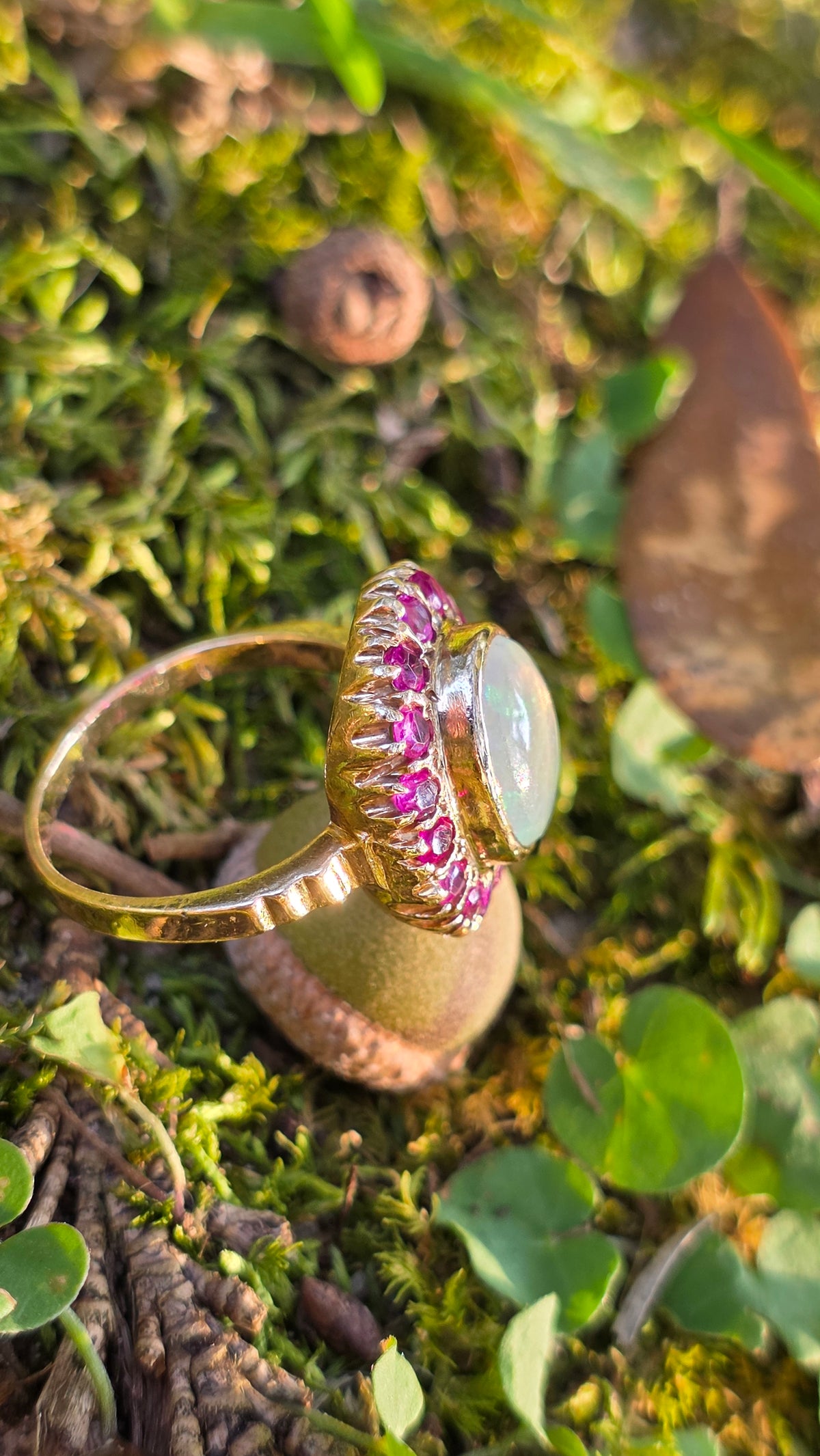 A gold ring with a large oval pale green stone and rows of pink gemstones, photographed resting on moss and small green leaves outdoors.